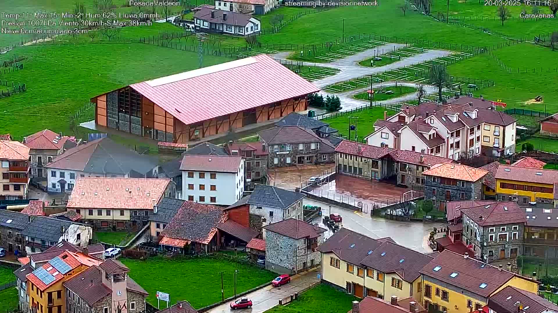 An aerial view shows a small village with stone and tile-roofed buildings, a large reddish-brown building, and a wet paved area, all under overcast skies.
