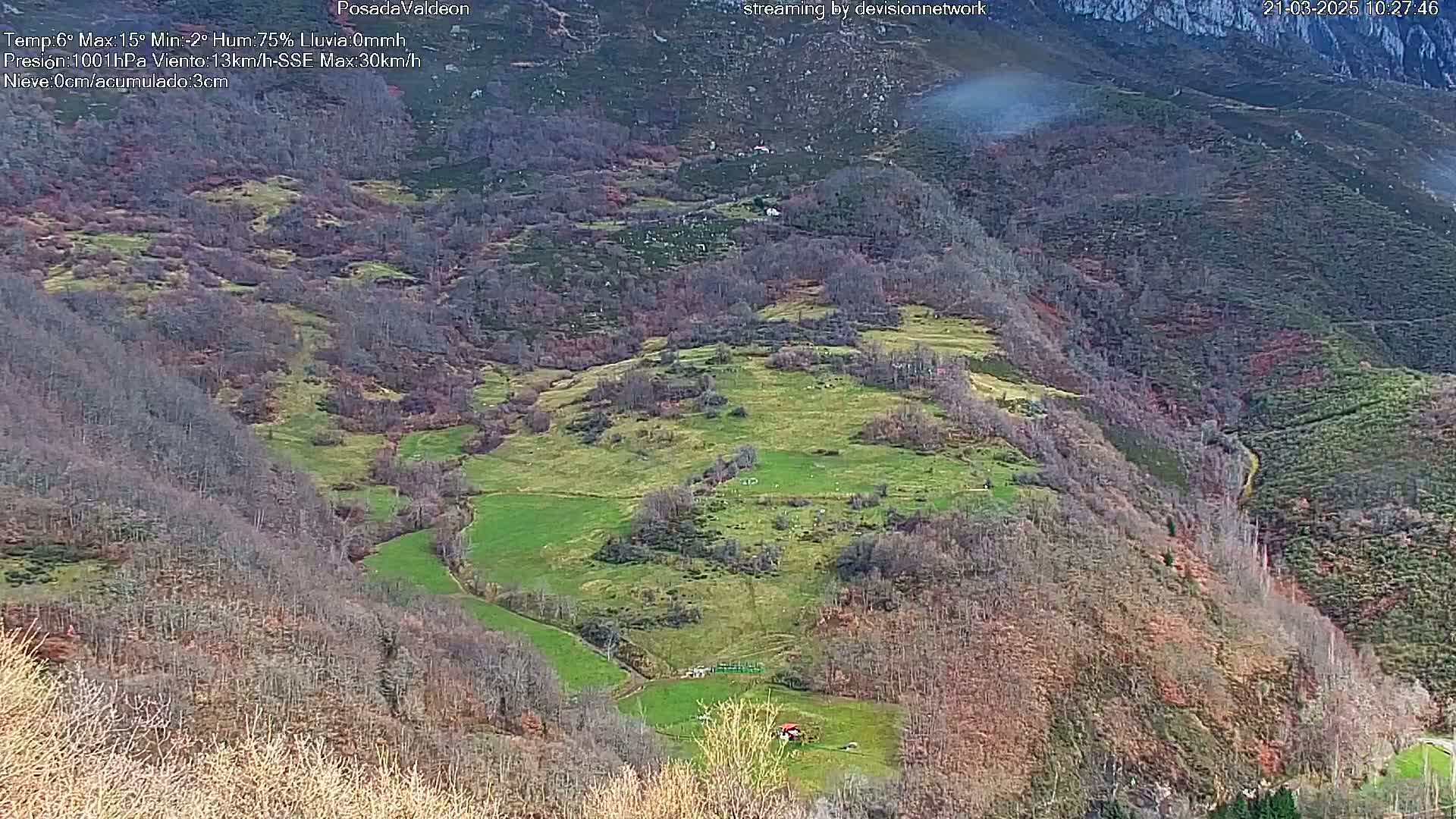 A high-angle view of a valley with patches of green pastureland, interspersed with leafless trees and shrubs, under a partly cloudy sky.
