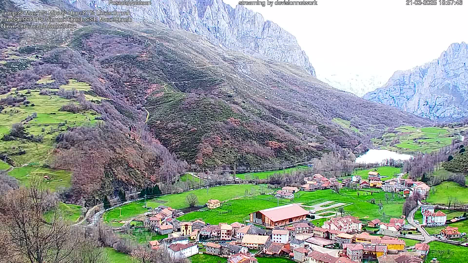 A small village nestled in a verdant valley between towering mountains, under a mostly cloudy sky.