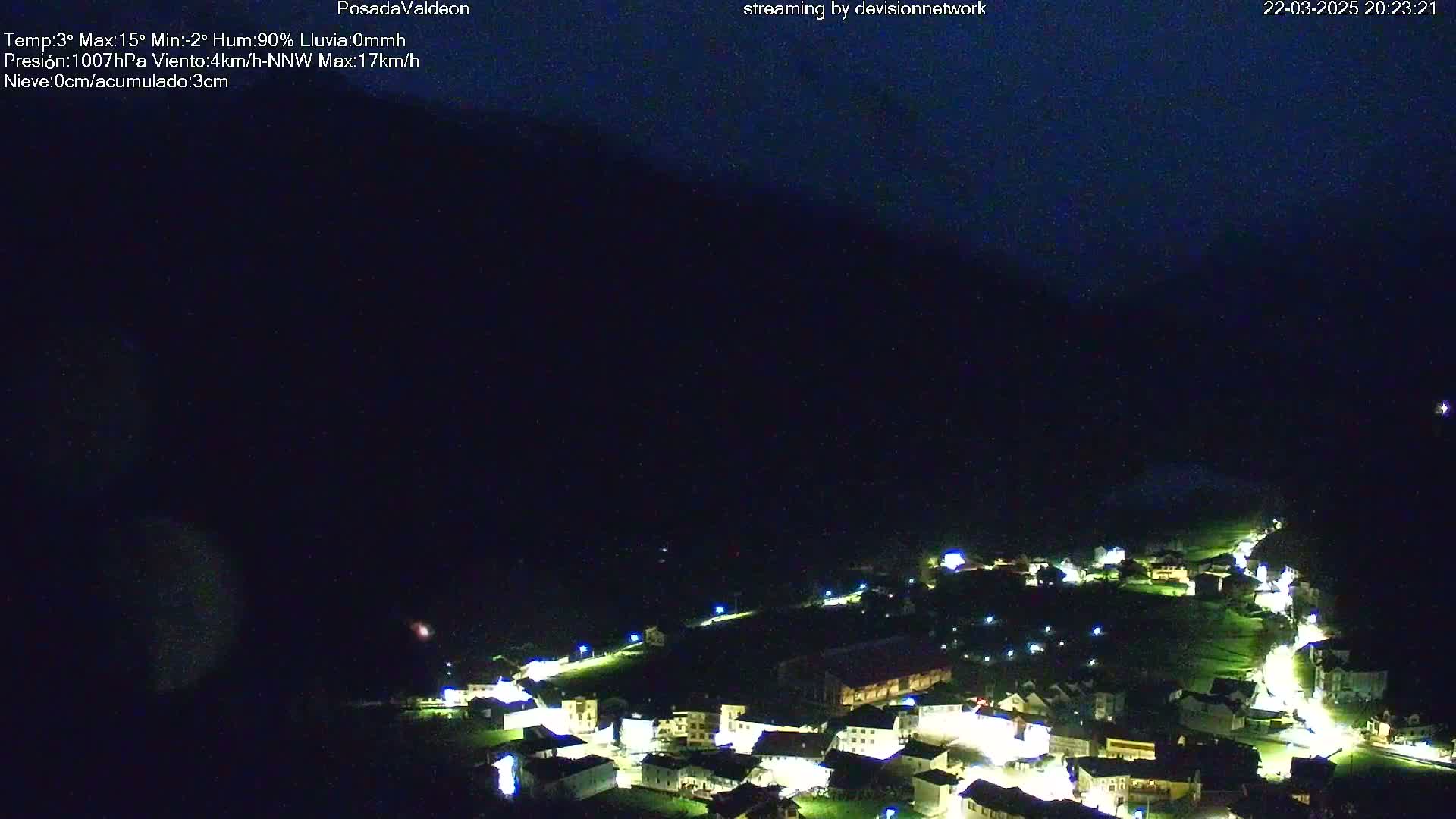 A nighttime aerial view shows a small village illuminated with lights nestled in a dark valley under a mostly clear, dark sky.