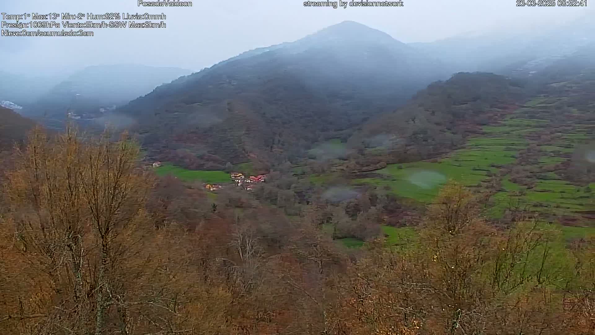 A misty, rainy view of a valley with terraced fields and a small village nestled among bare trees and rolling hills.
