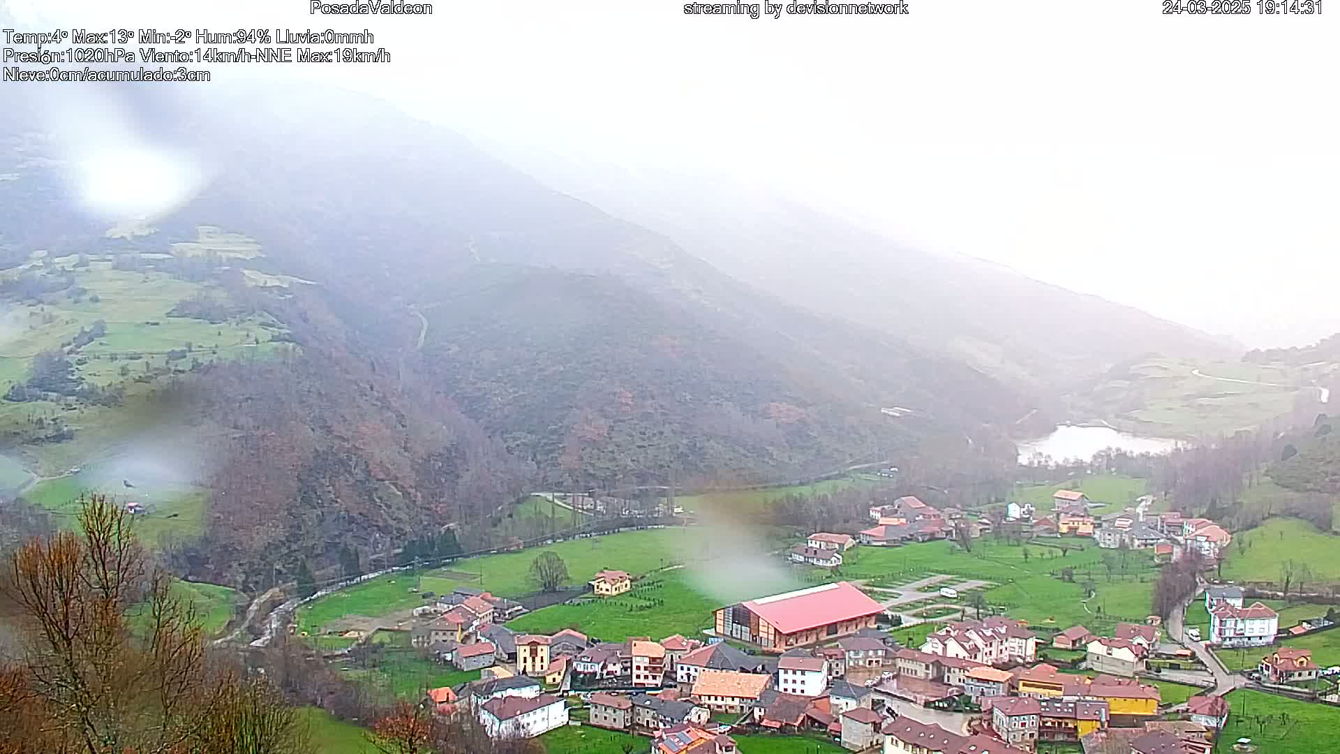 A small village nestled in a valley surrounded by mist-covered mountains, with a body of water visible in the distance.