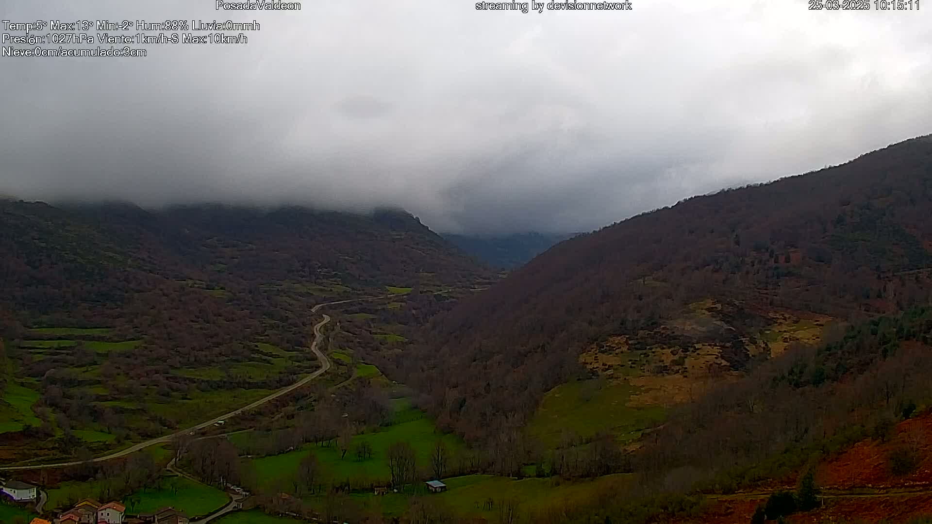 A winding road runs through a valley nestled between misty, tree-covered mountains under a cloudy sky.