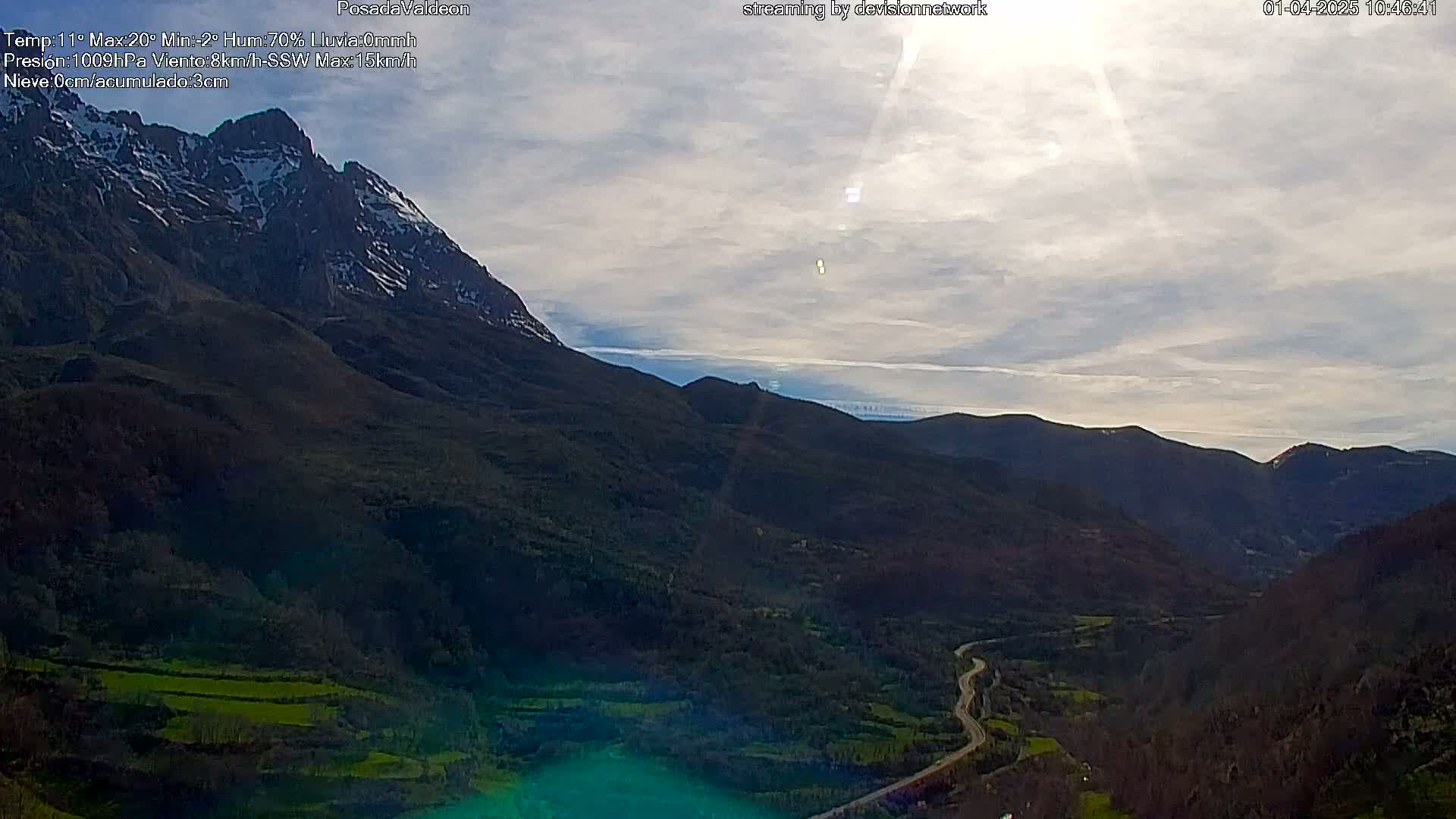 A mountain range with a snow-capped peak is visible under a partly cloudy sky, with a winding road snaking through a valley below.