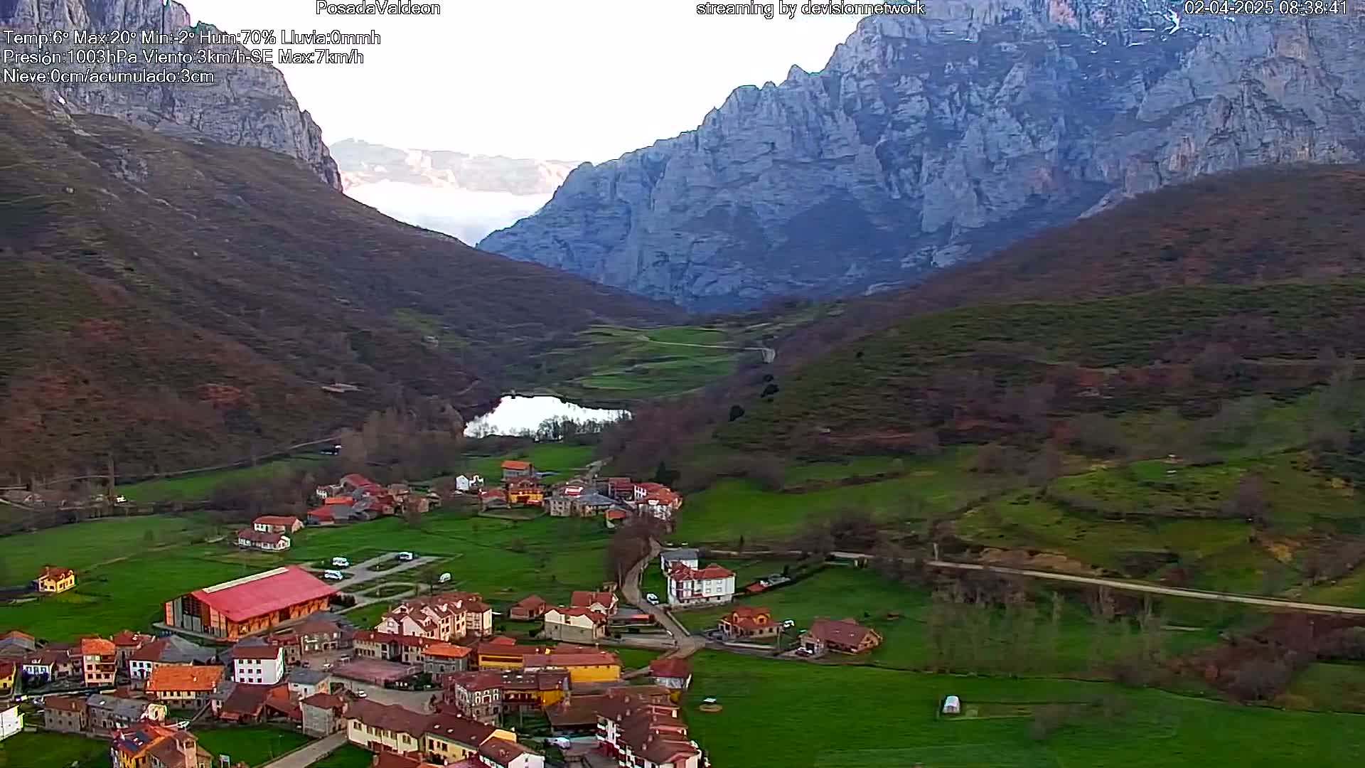 An aerial view of a small village nestled in a valley between large, grey mountains, under partly cloudy skies.