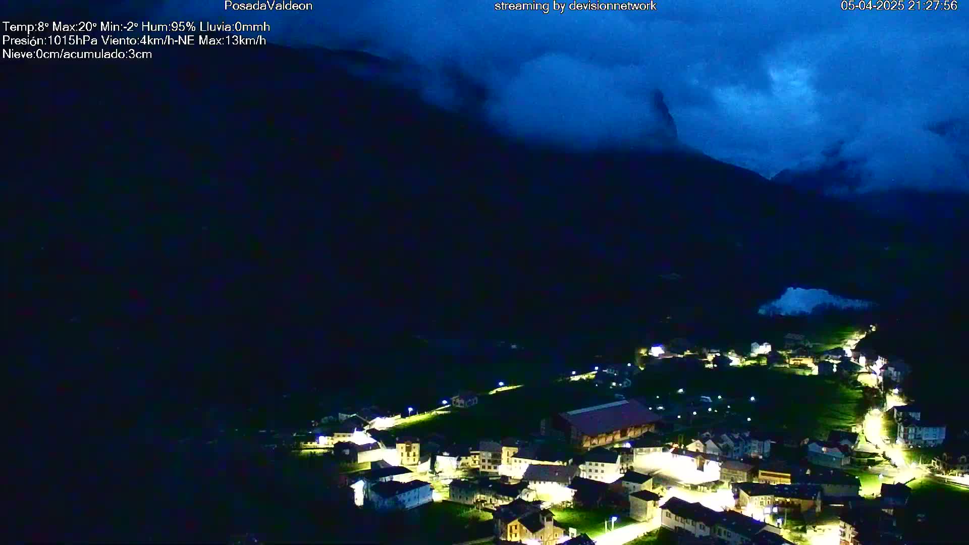 A nighttime view of a small village nestled in a valley, illuminated by streetlights, under a dark, cloudy sky.