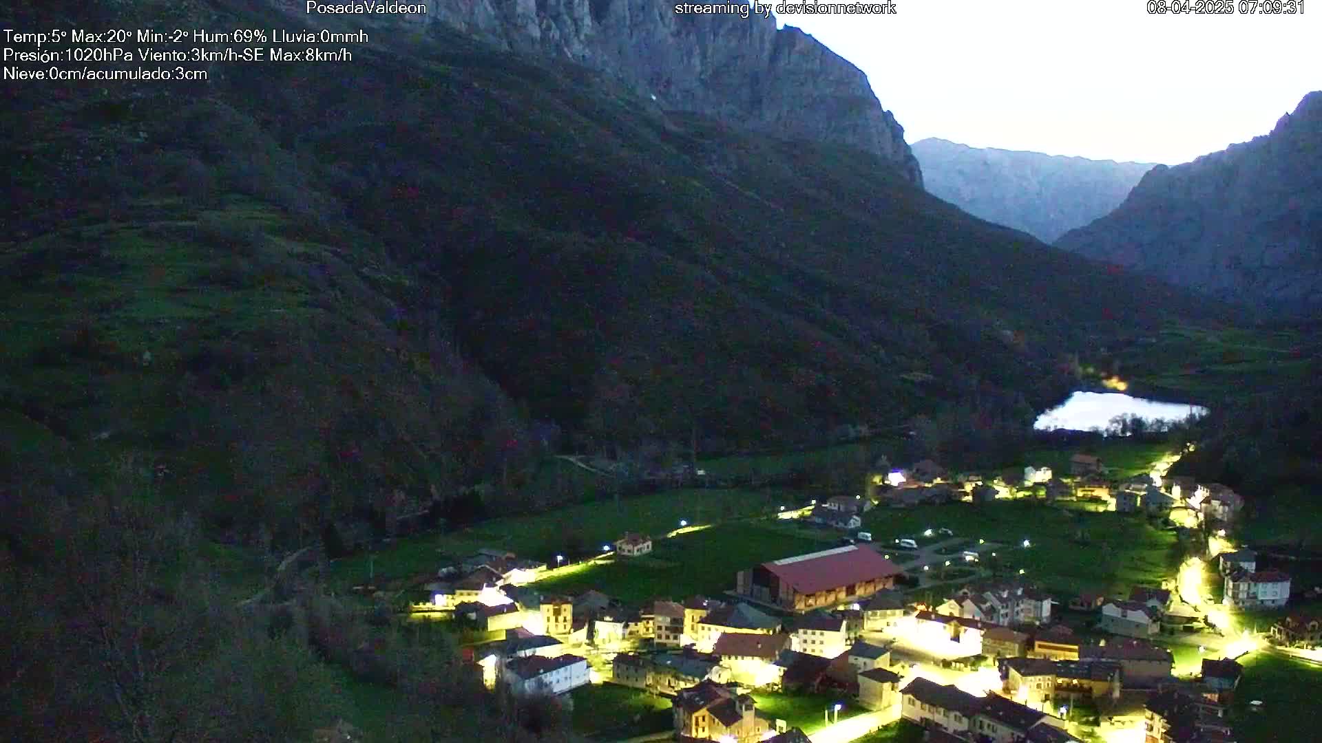 A small village nestled in a valley between dark mountains is illuminated at night under a twilight sky.