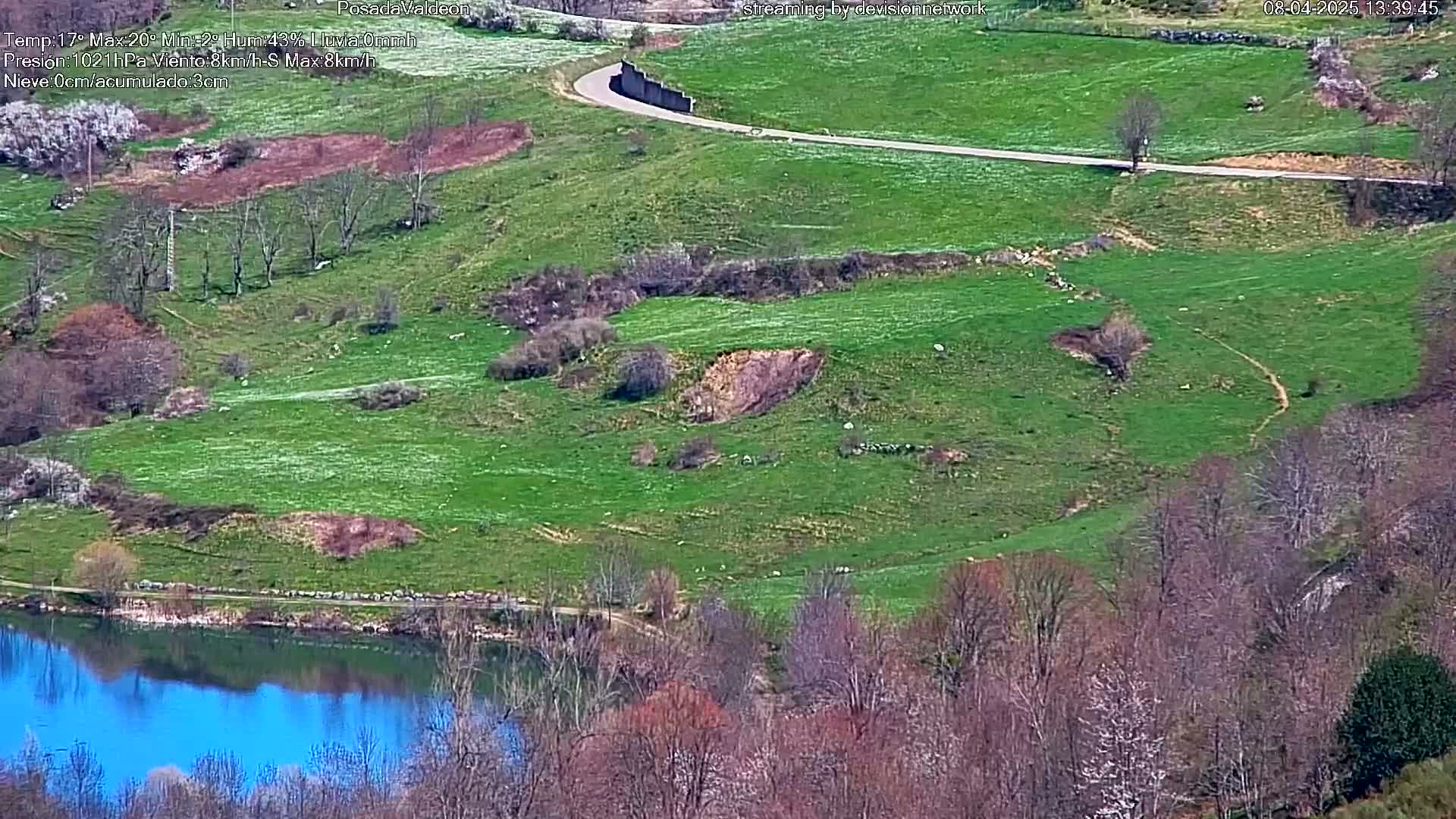A winding road curves through a verdant hillside dotted with sparse trees and small, barren patches of earth, leading to a calm blue lake reflecting the surrounding landscape.