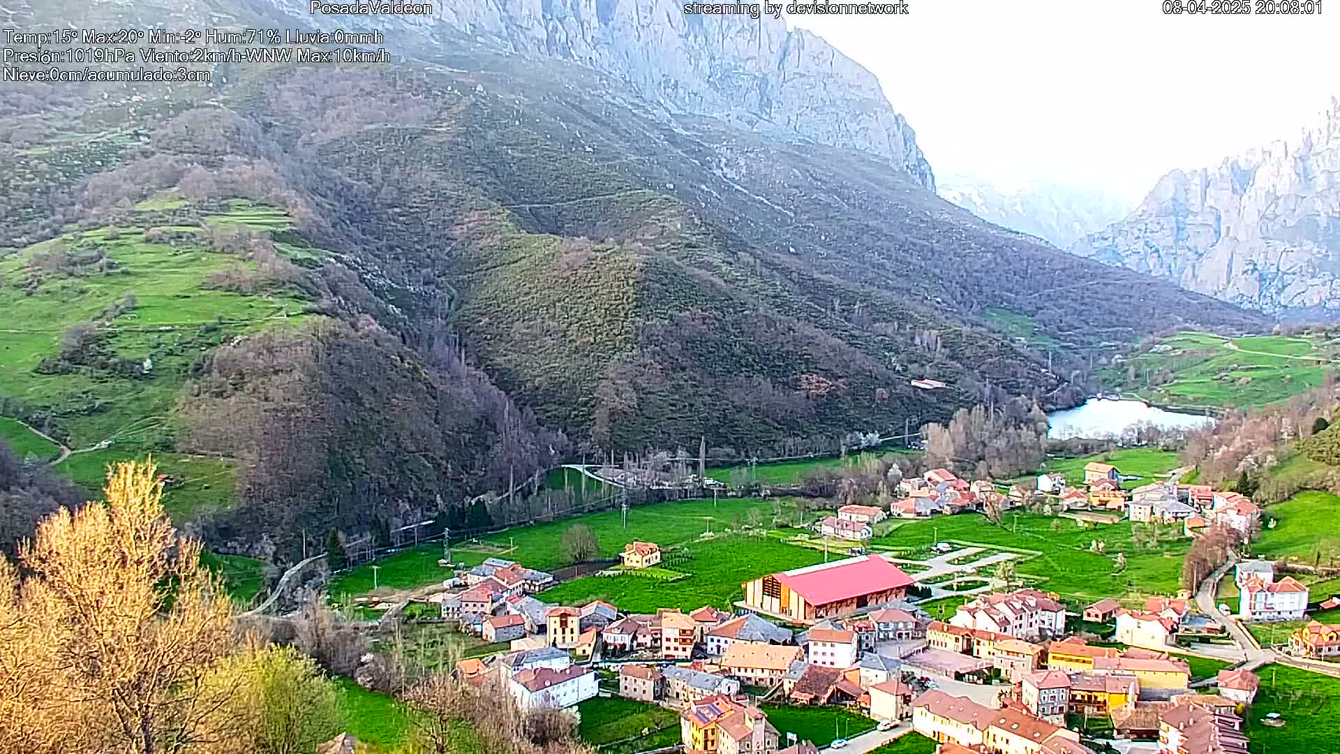 A small village nestled in a verdant valley between towering mountains, under a partly cloudy sky.