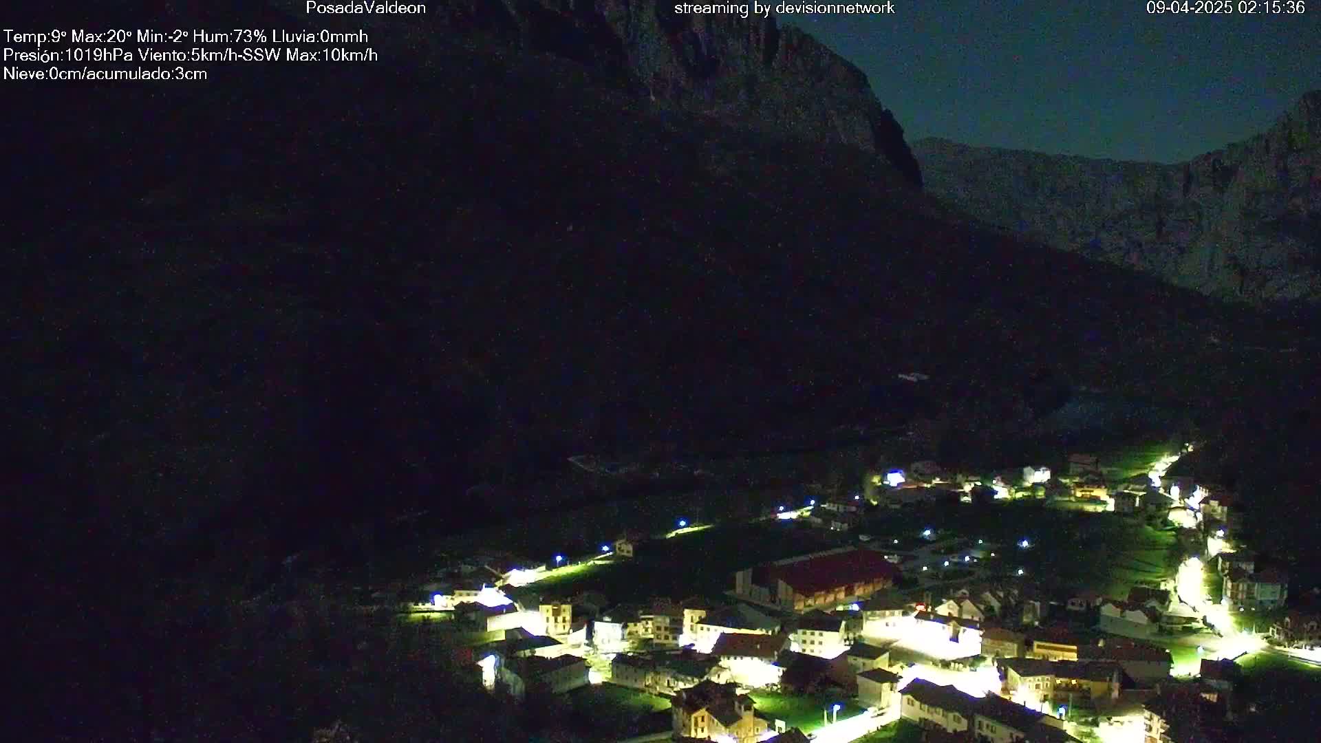 A small village is illuminated at night under a dark, clear sky nestled against a large, dark mountain.