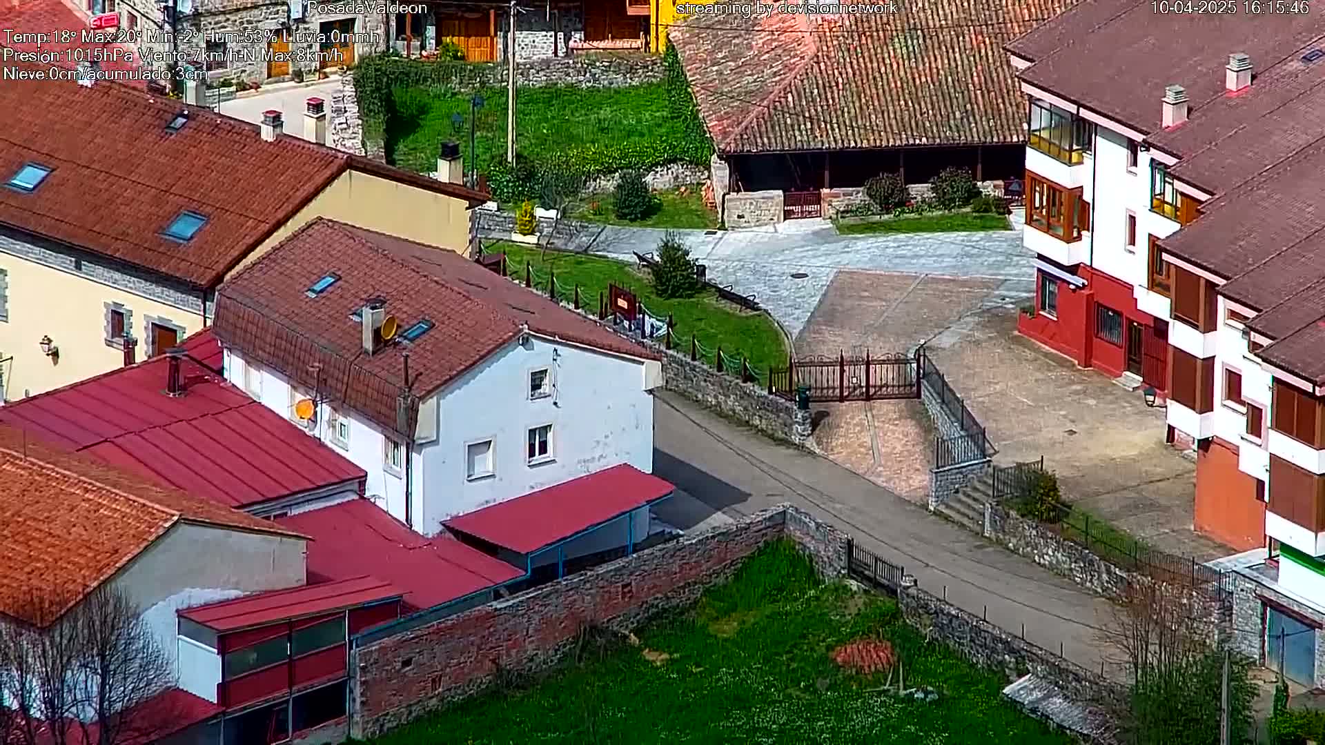 A high-angle view of a village on a sunny day shows red-tiled rooftops, white-walled buildings, and grassy areas.