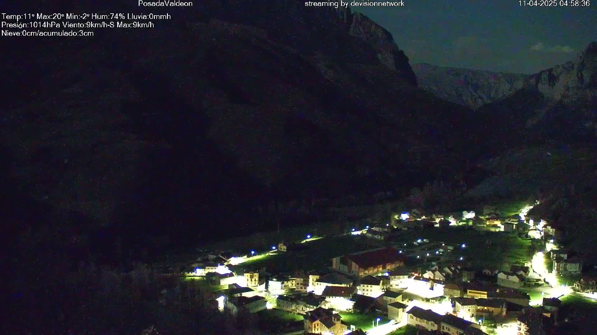 A nighttime view of a small, illuminated village nestled in a dark valley beneath a large, shadowed mountain under a clear night sky.