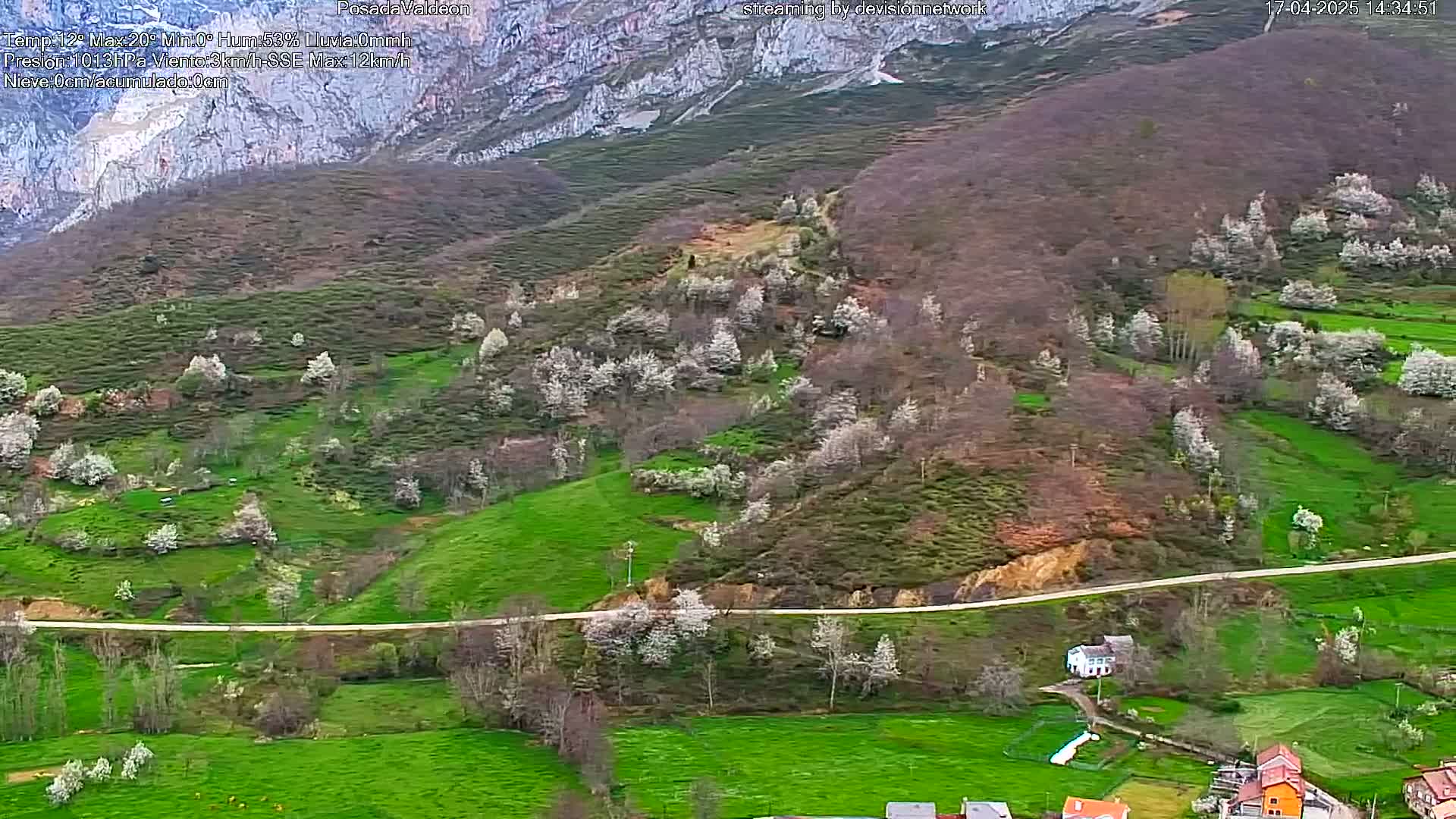 A hilly landscape with green fields and blossoming trees is seen from above, a road curves through the scene, and a few buildings are visible near the bottom.
