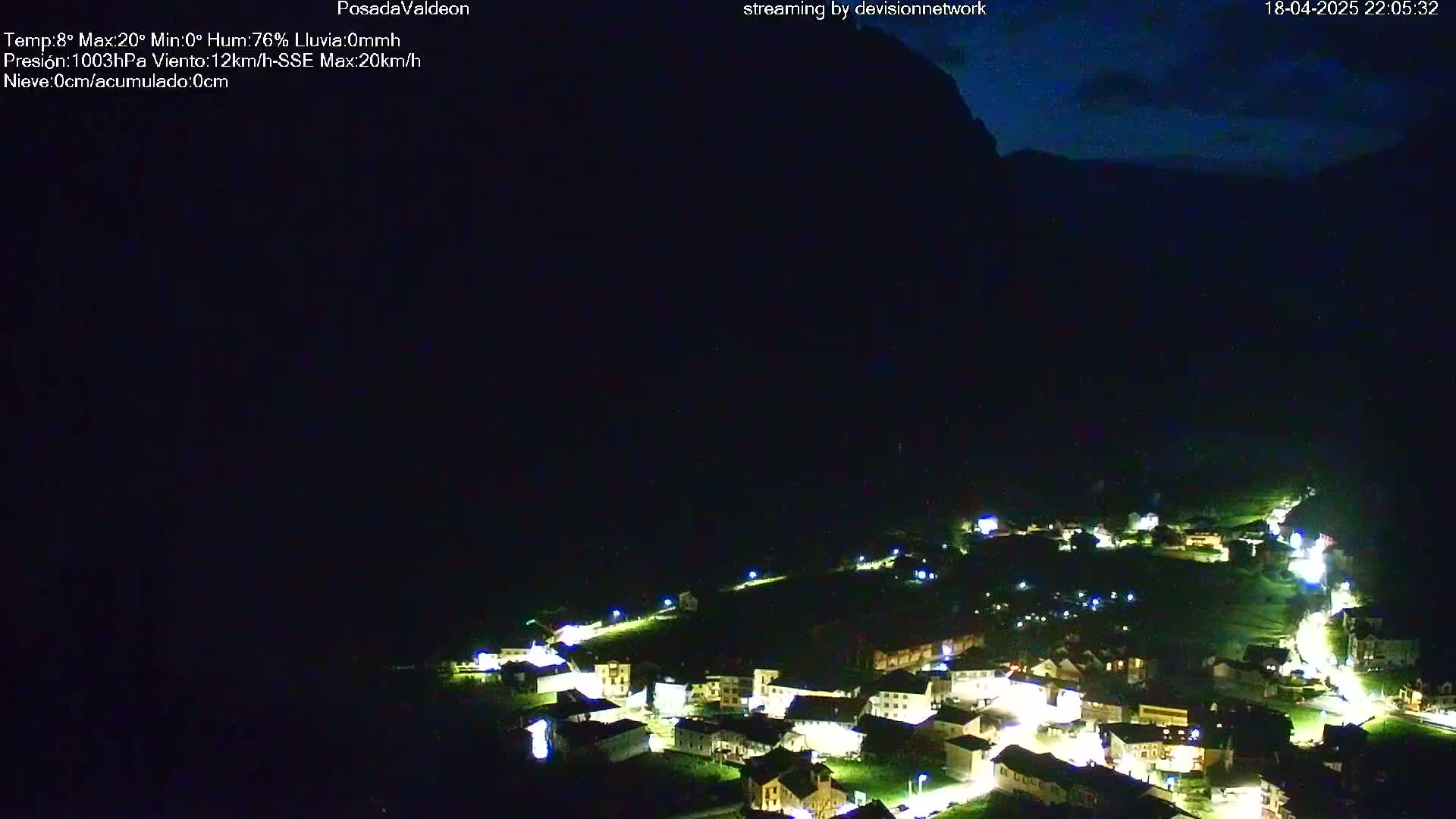 At night, a small village with illuminated buildings is nestled in a dark valley under a mostly clear, dark blue sky.