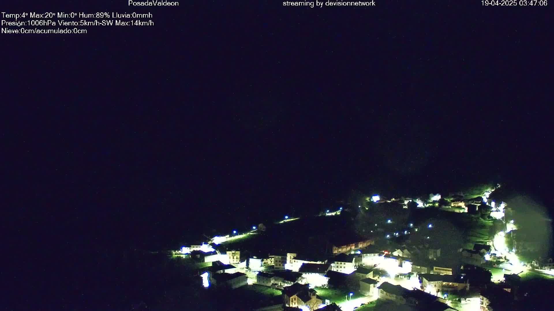 A nighttime aerial view shows a small village illuminated by lights nestled on a hillside under a dark, clear sky.