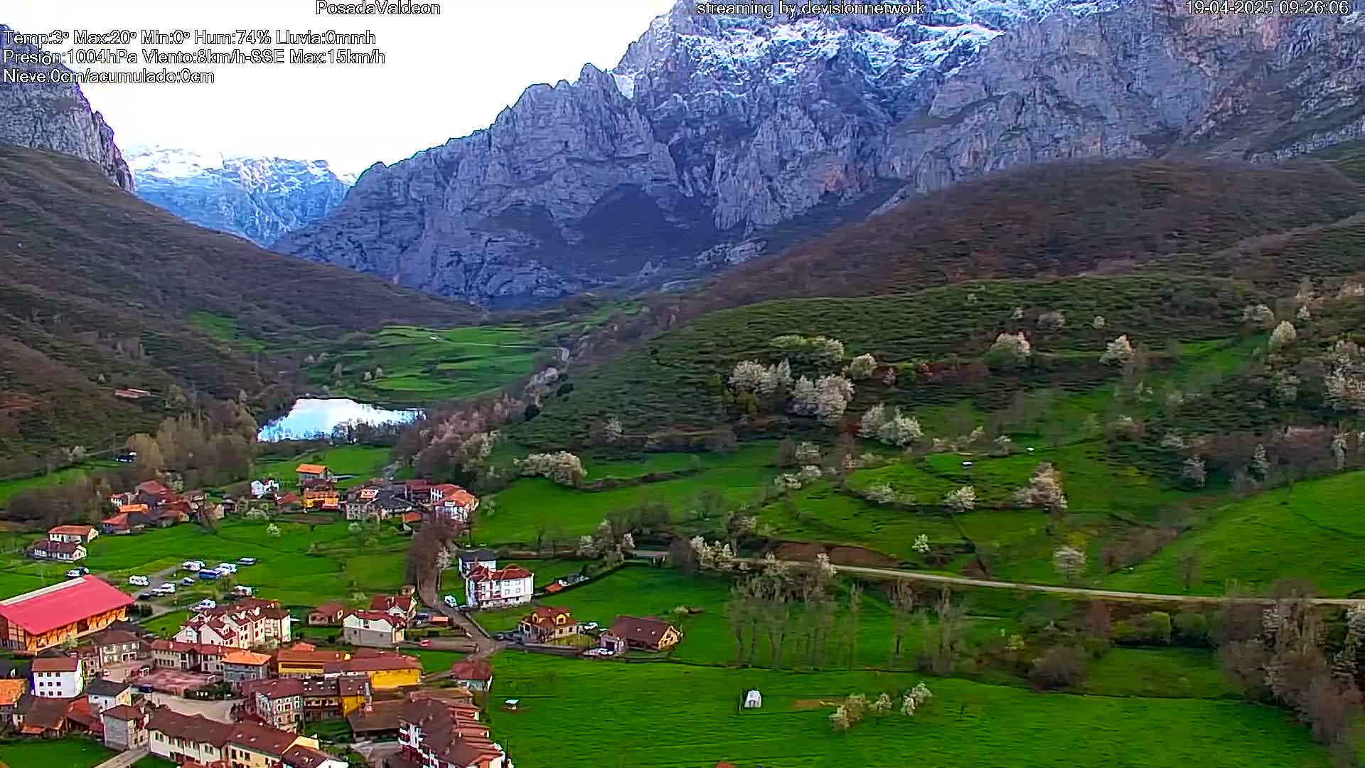 An aerial view shows a small village nestled in a green valley, with snow-capped mountains in the background under partly cloudy skies.