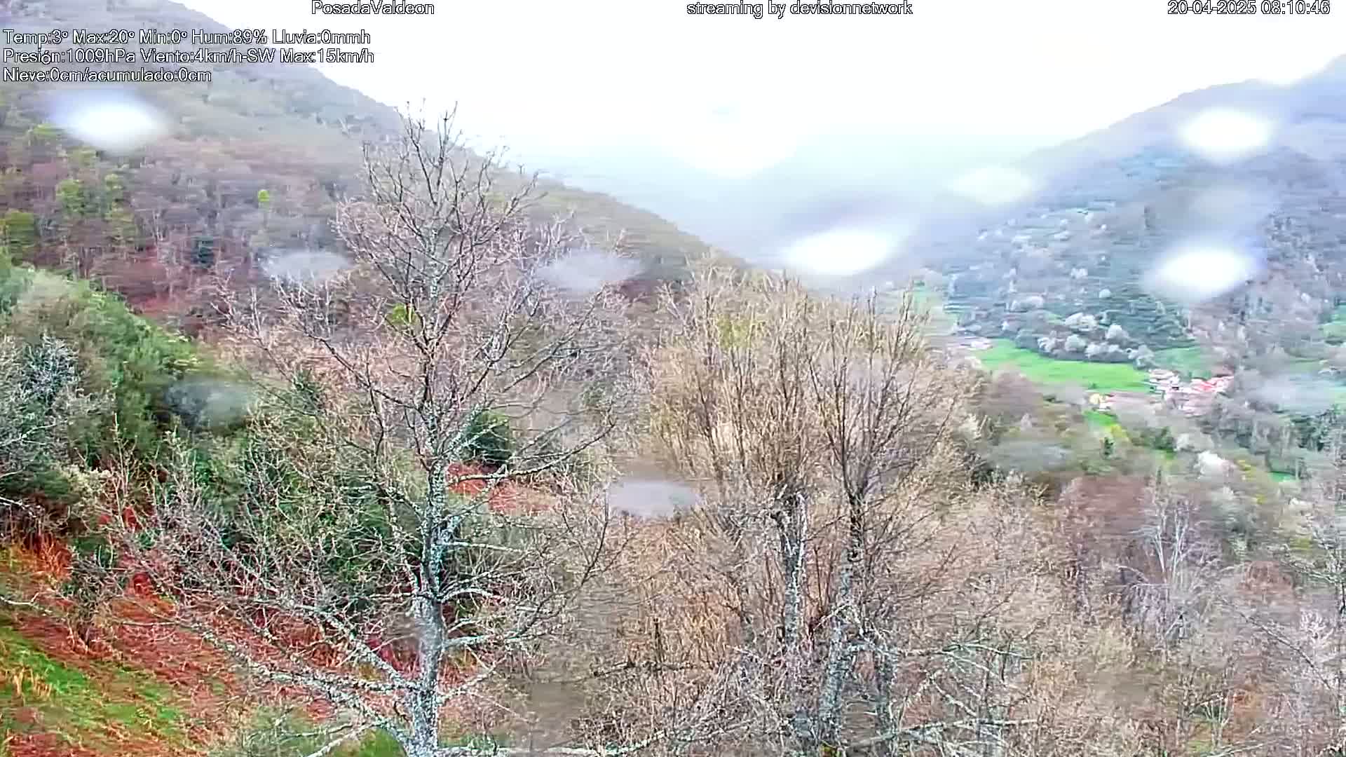 A rain-swept view of a valley dotted with leafless trees and small buildings, nestled among hillsides.