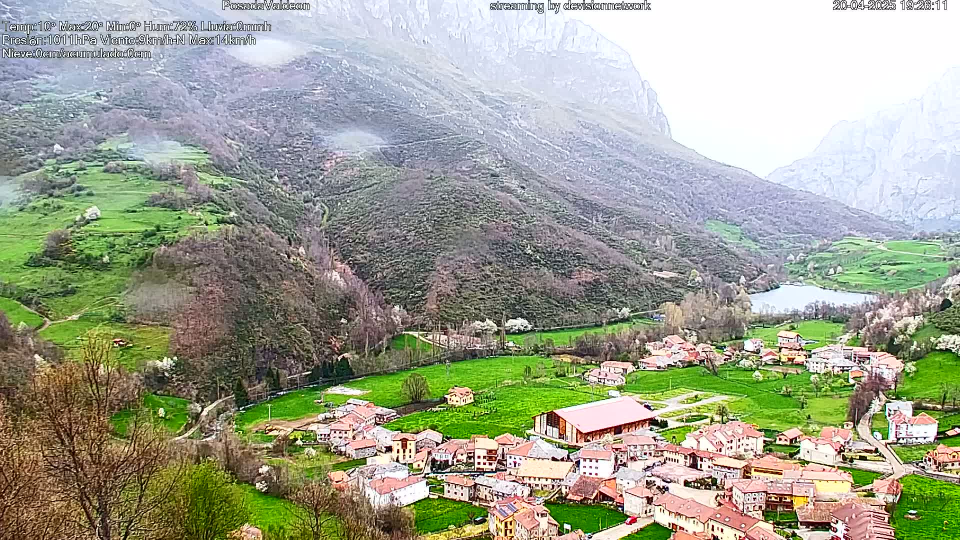 A small village nestled in a valley between mountains on an overcast day, with green fields and a lake visible.