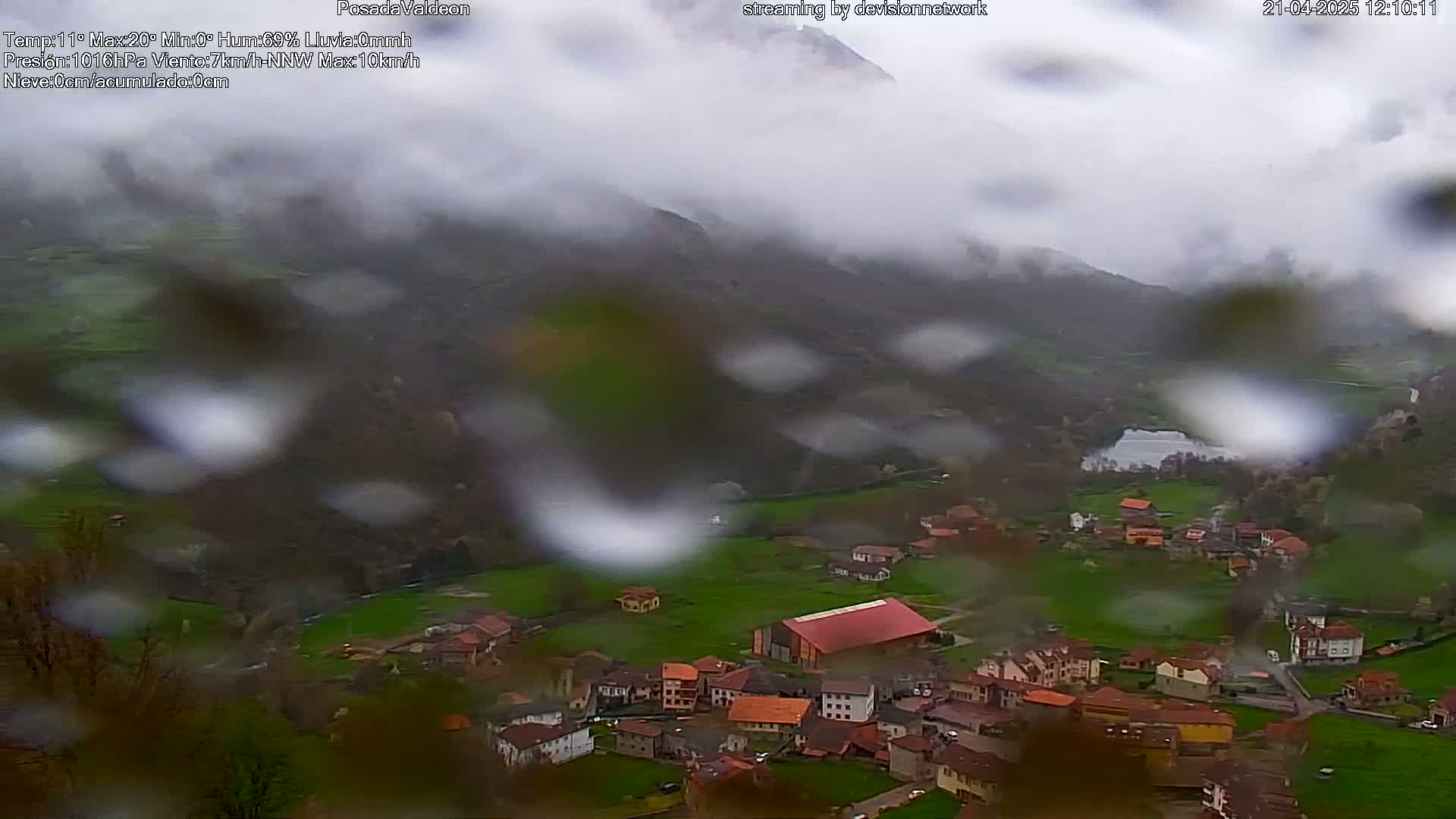 Rain falls on a small village nestled in a valley between green hills and mountains shrouded in fog.