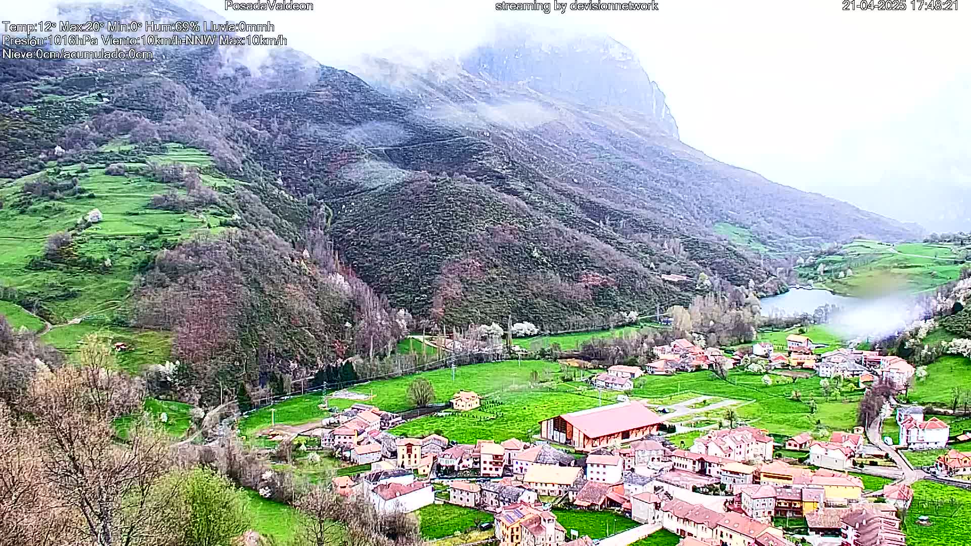 A misty valley village nestled between green hills and a large mountain, under a cloudy sky.