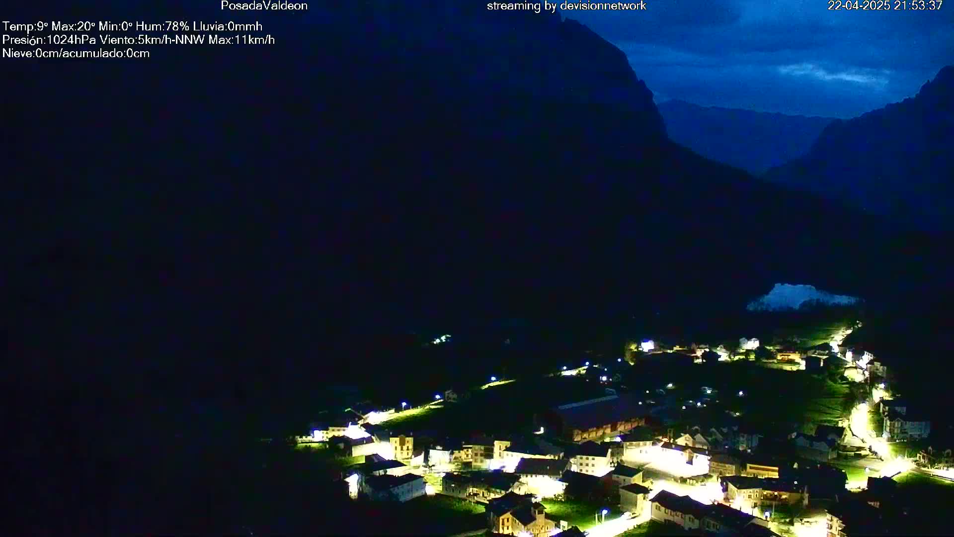 A nighttime view of a small village nestled in a valley between dark mountains under a twilight sky.
