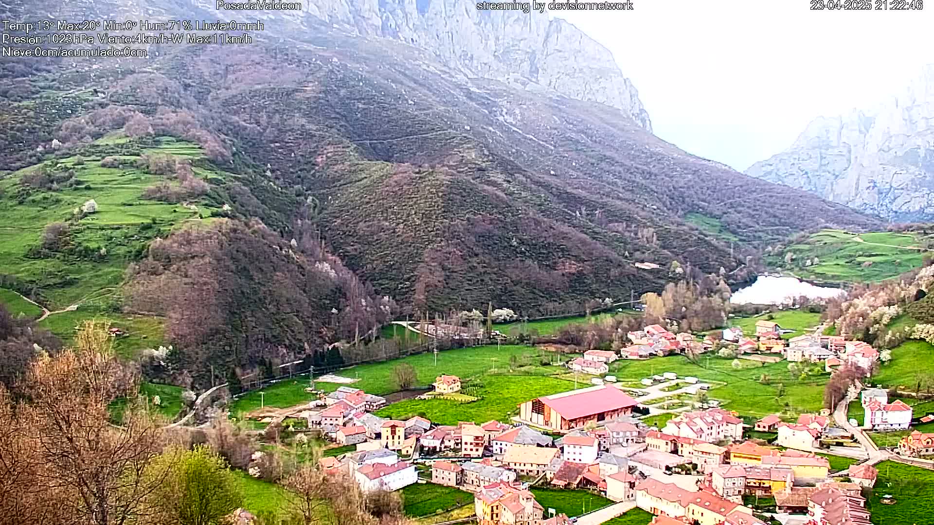A small village nestled in a valley between mountains, with green fields and a calm body of water visible in the distance, under an overcast sky.