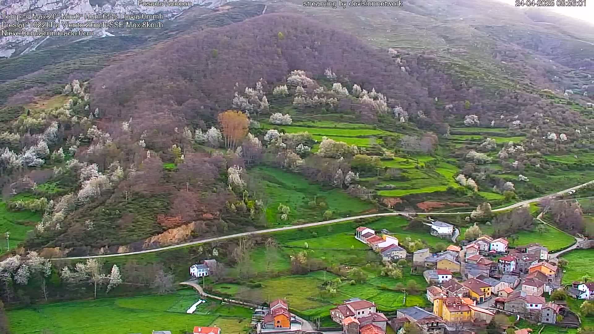 An aerial view shows a small village nestled in a verdant valley surrounded by hillsides dotted with blossoming trees under a partly cloudy sky.