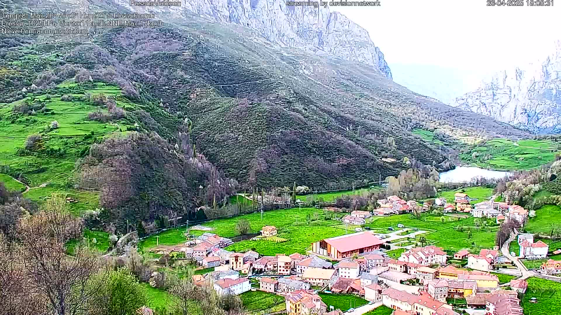 A small village nestled in a verdant valley between steep, rocky mountains, under a mostly cloudy sky.
