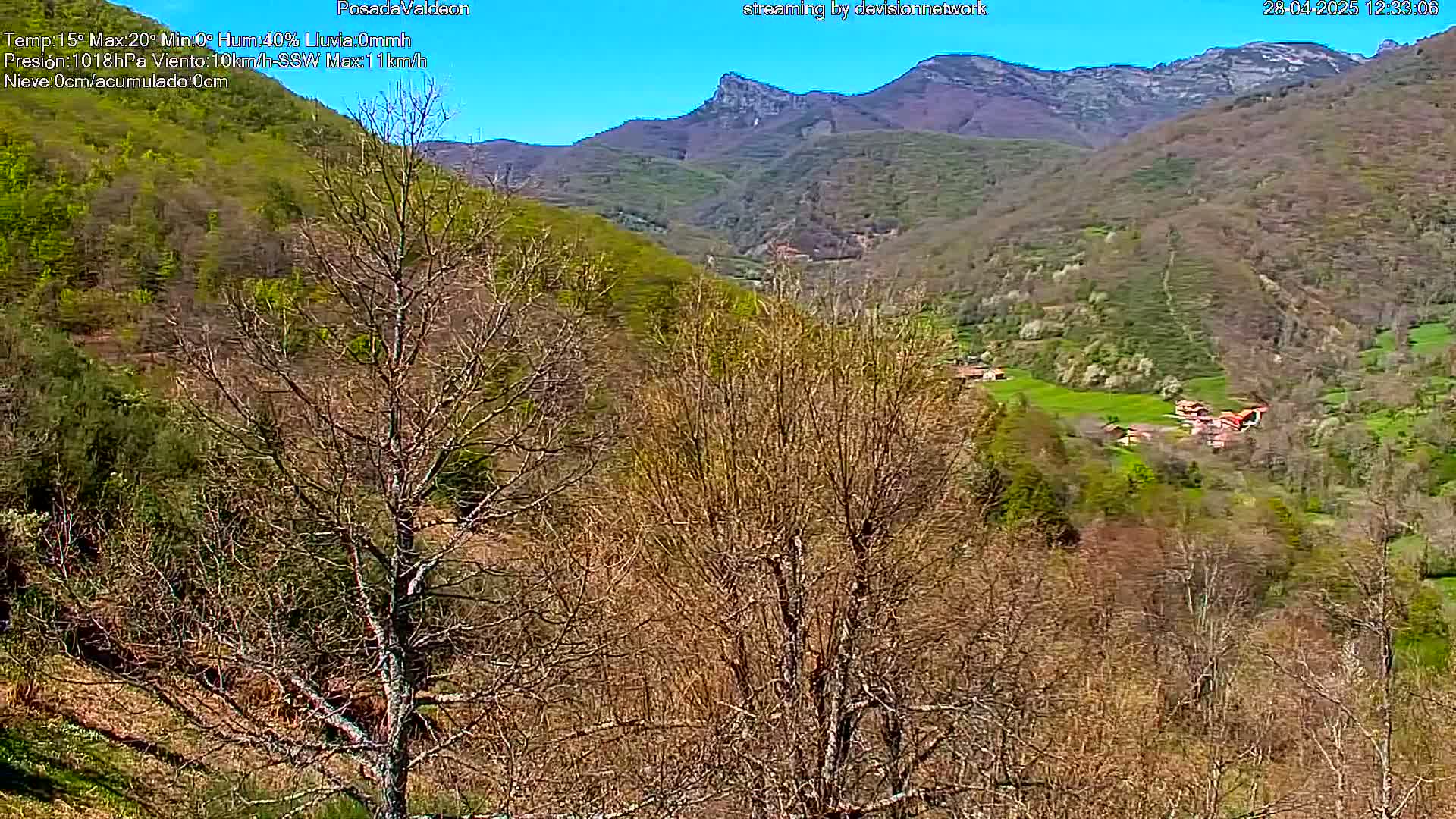 A high-angle, long shot reveals a valley scene under a clear blue sky;  leafless trees in the foreground contrast with budding vegetation and a small village nestled in a valley surrounded by hills.