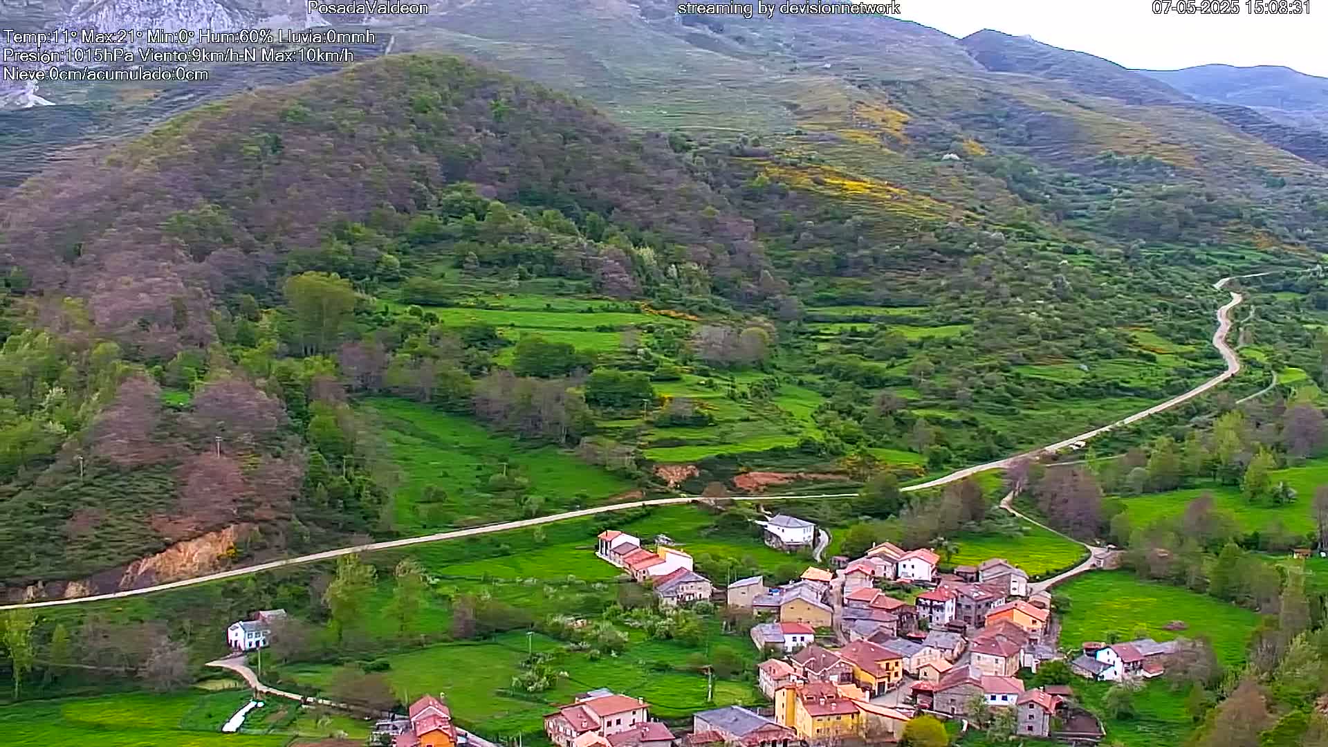 An aerial view shows a small village nestled in a verdant valley, with a winding road leading up to it, all under an overcast sky.