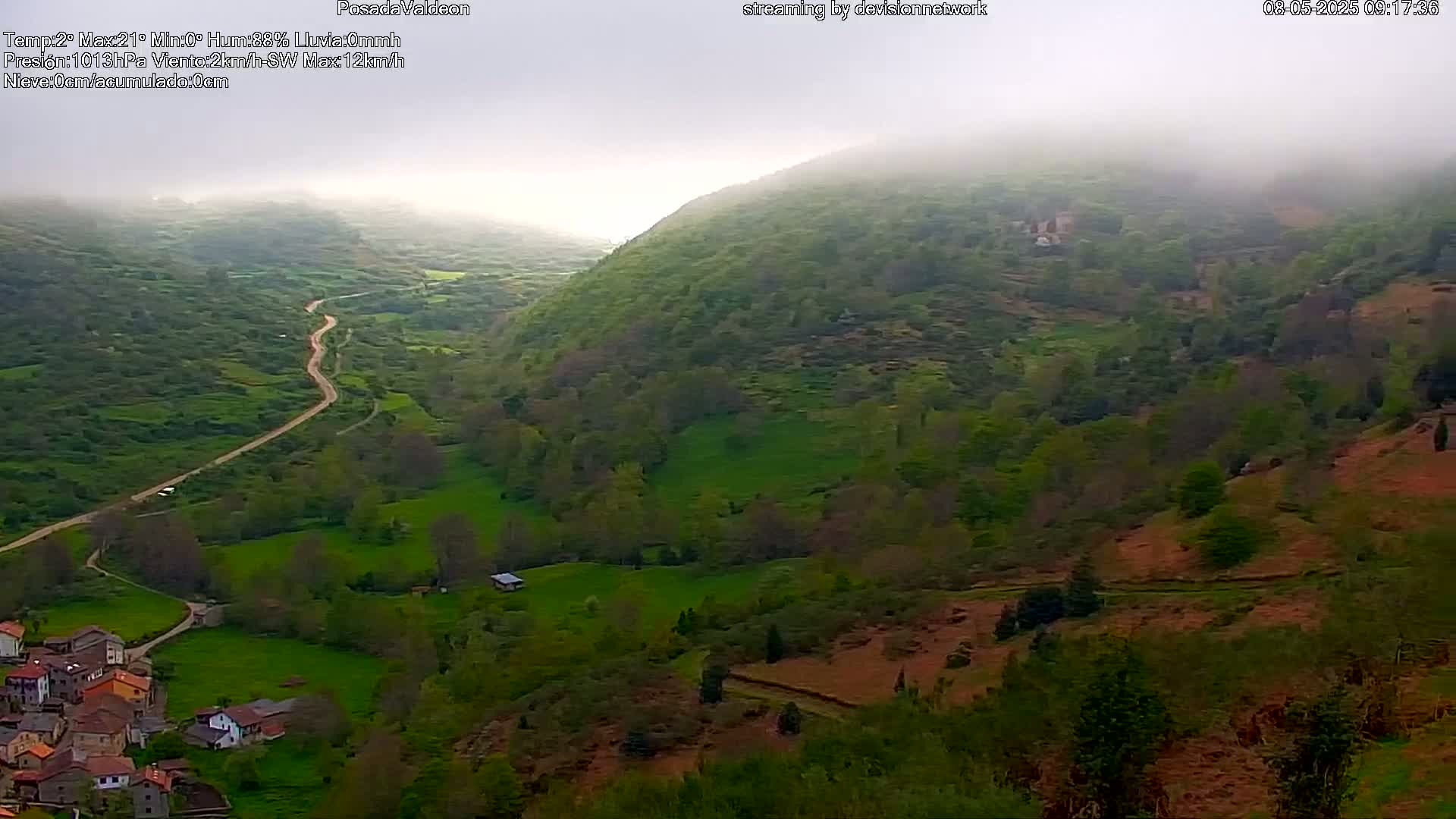 A winding road leads through a misty green valley to a small village nestled below fog-covered hills.
