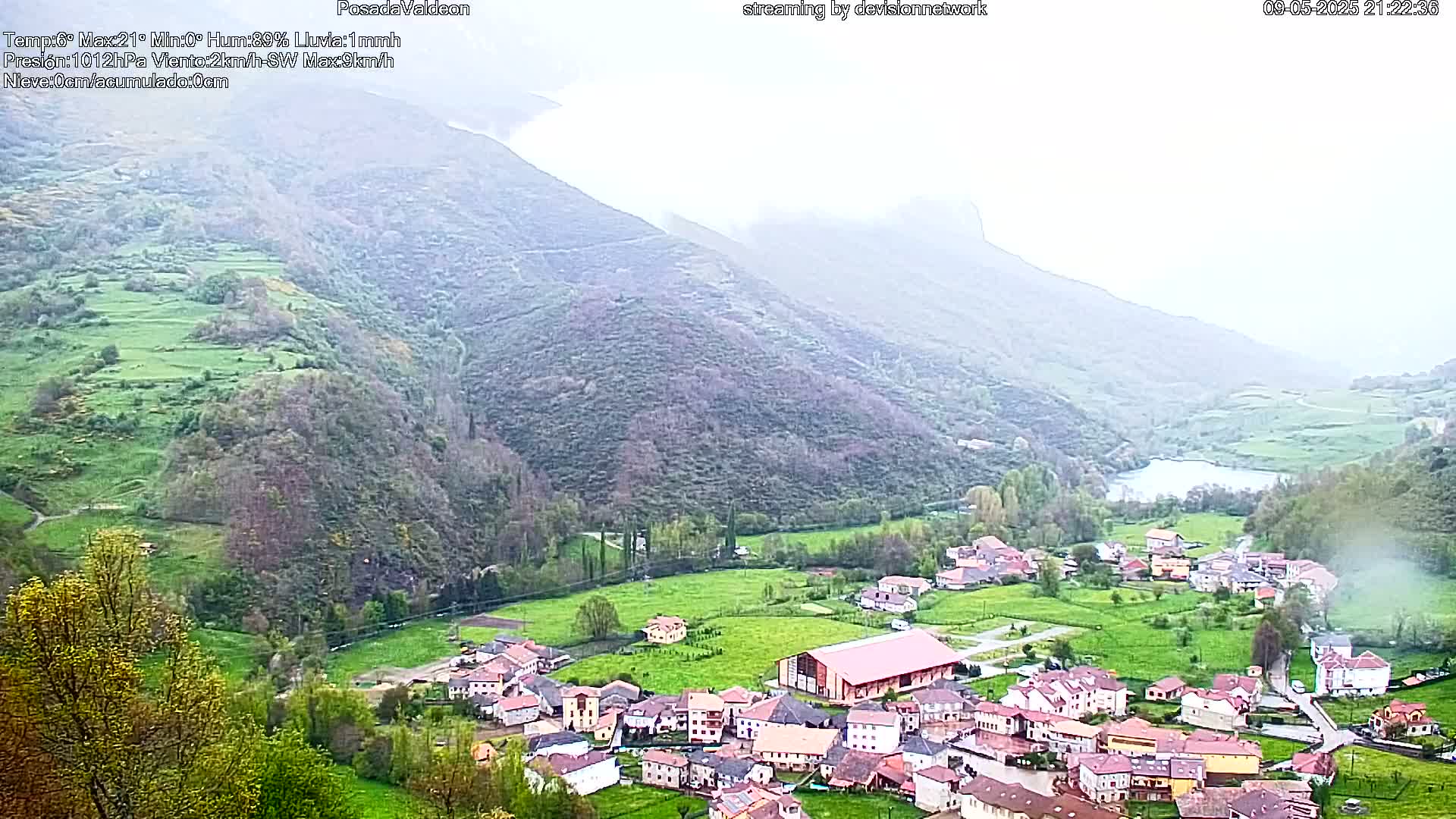 A small village nestled in a valley between misty green mountains, with a lake visible in the distance.