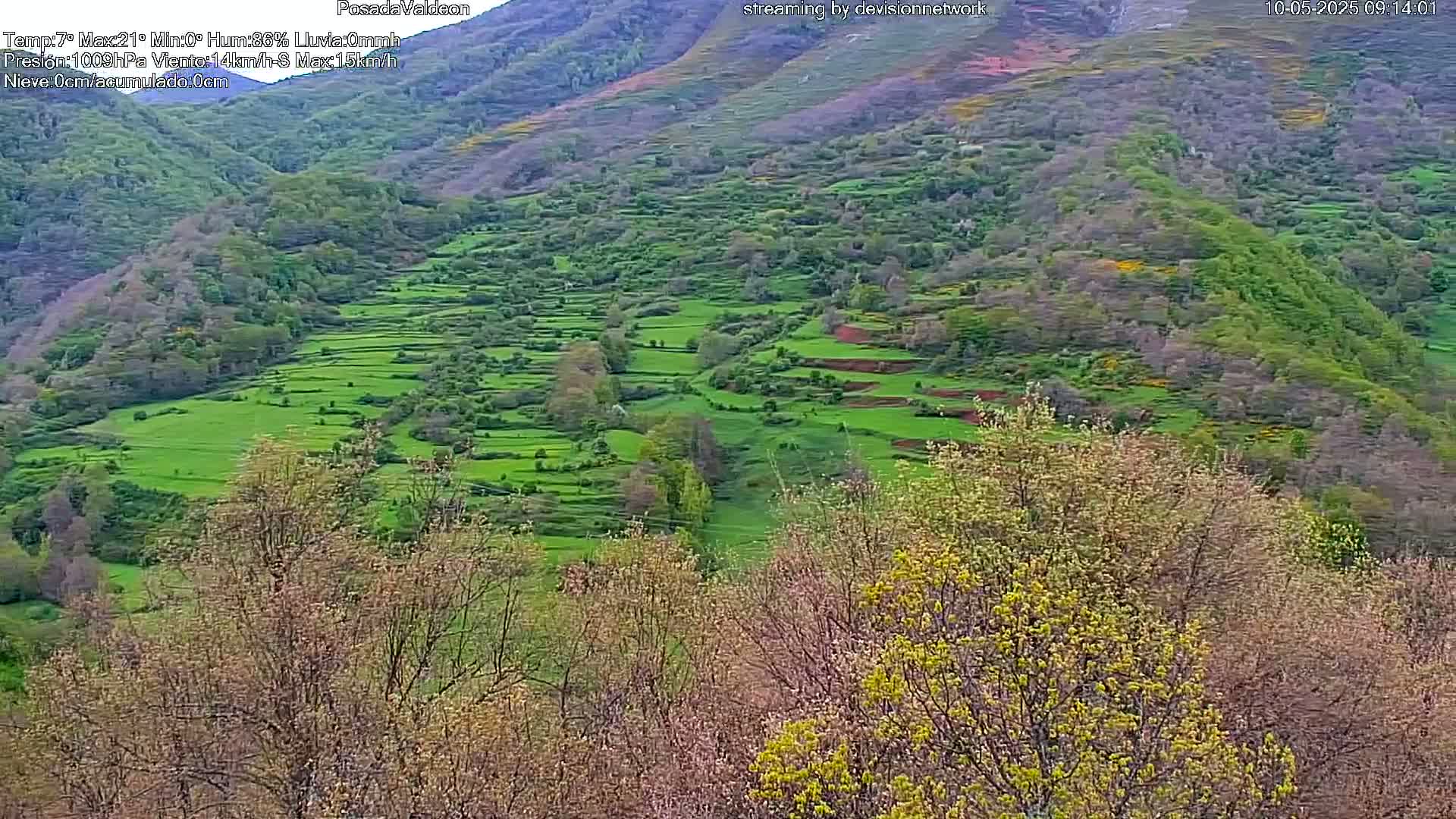 A hillside covered in terraced green fields and budding trees is visible under a partly cloudy sky.