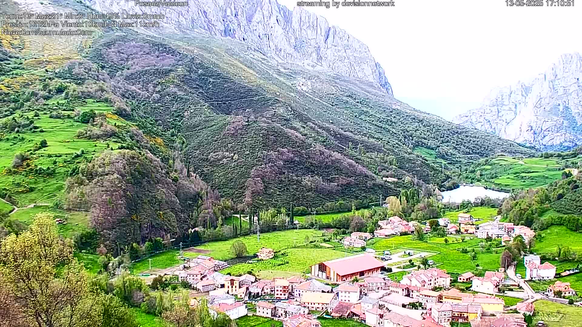 A small village nestled in a verdant valley between towering, rocky mountains, under a partly cloudy sky.