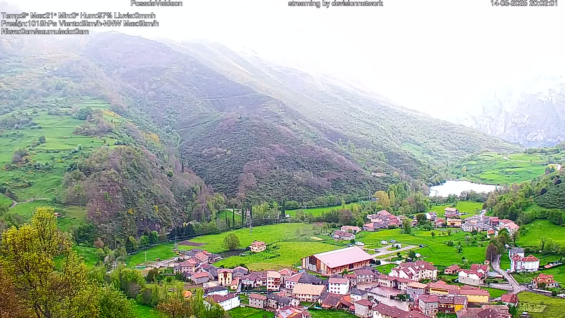 A small village nestled in a green valley, surrounded by hills and a small lake, under overcast skies.