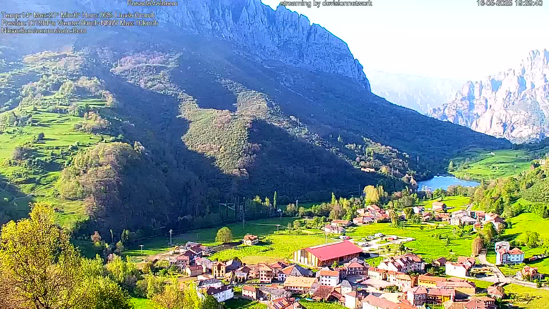 A sunny vista shows a small village nestled in a verdant valley between towering, rocky mountains, with a calm body of water visible in the valley.
