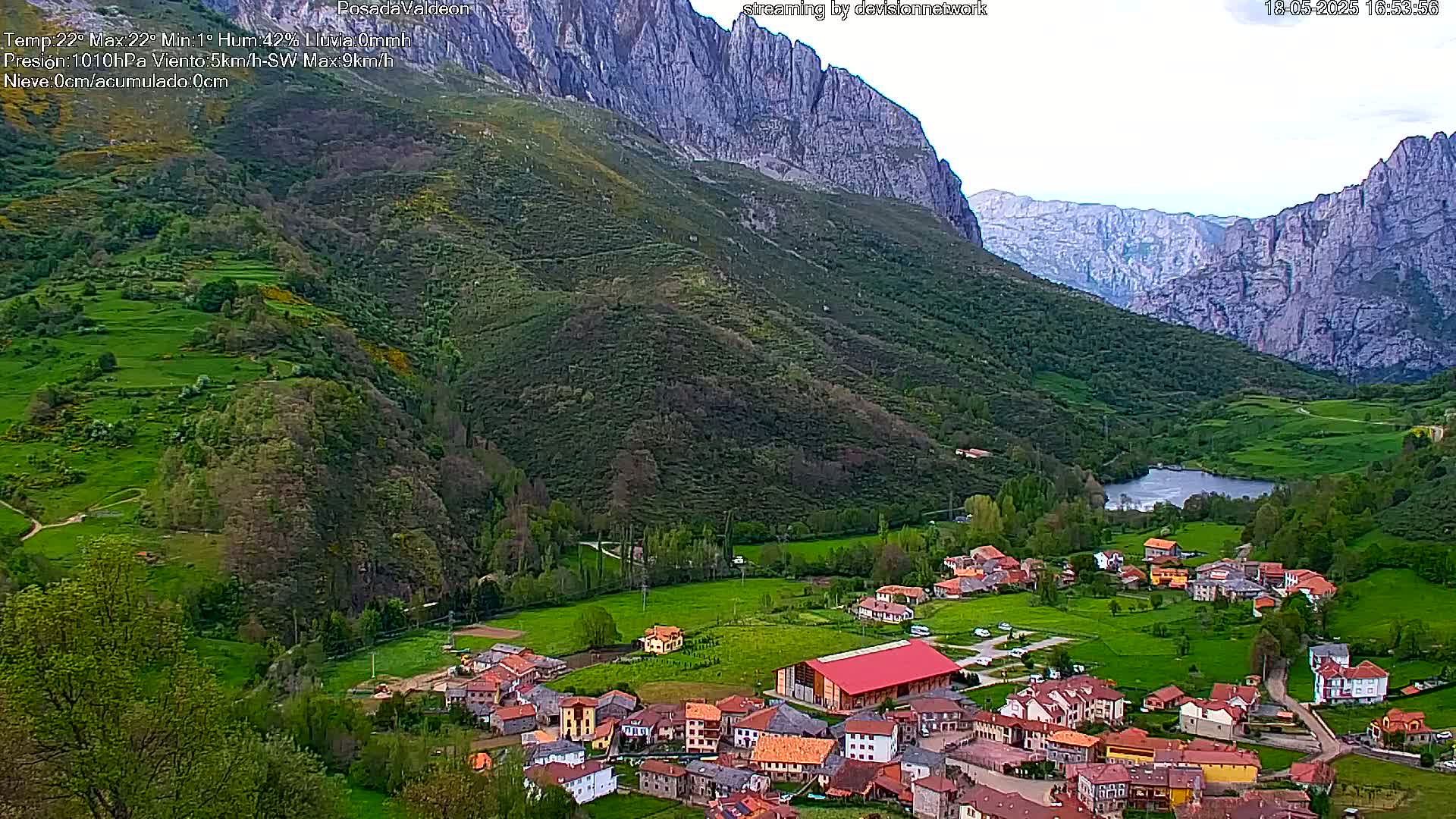 A small village nestled in a verdant valley between towering, light-grey mountains, under a partly cloudy sky.