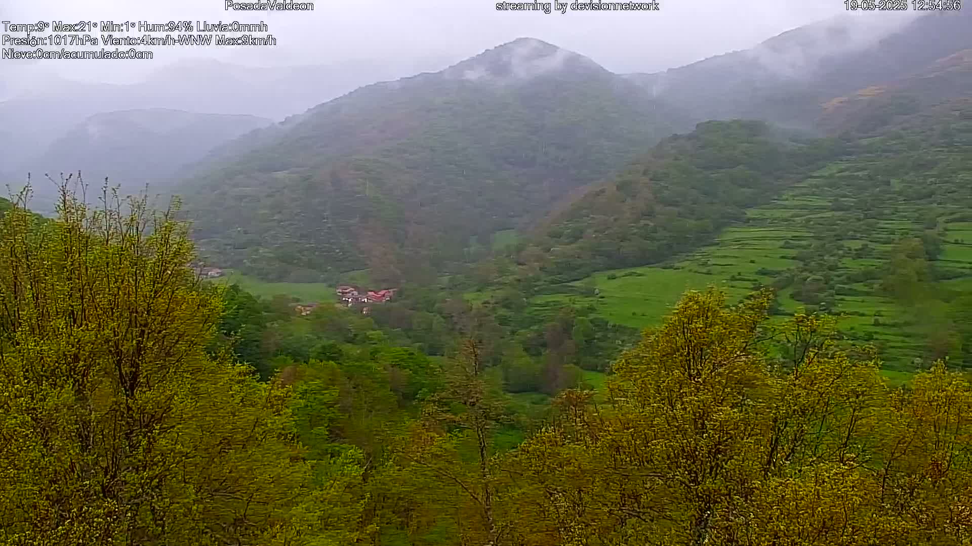 A misty, rainy view of a valley dotted with green fields and trees, nestled between rolling hills.