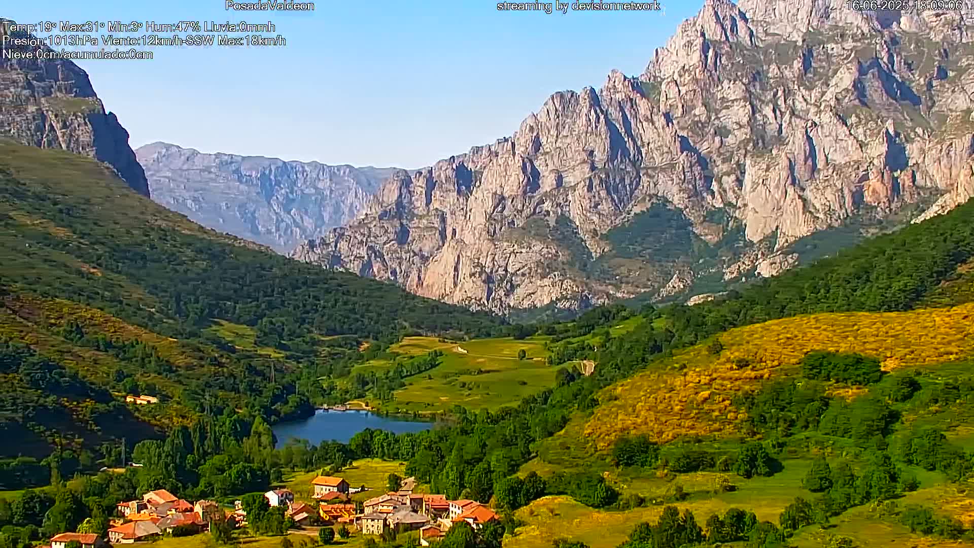A picturesque village nestled in a verdant valley, overlooked by towering, light-grey mountains under a clear, sunny sky.