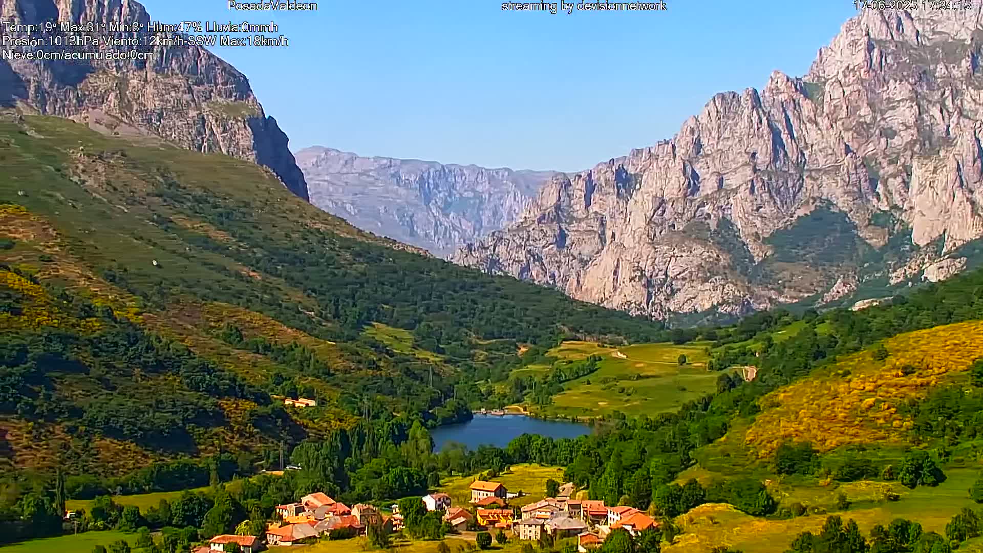 A small village nestled in a verdant valley between towering, rocky mountains under a clear blue sky.
