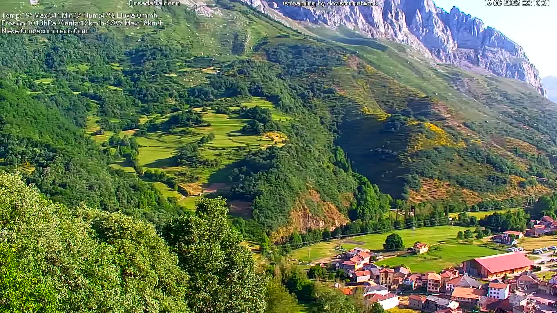 A picturesque village nestled in a verdant valley, surrounded by rolling green hills and mountains under a sunny sky.
