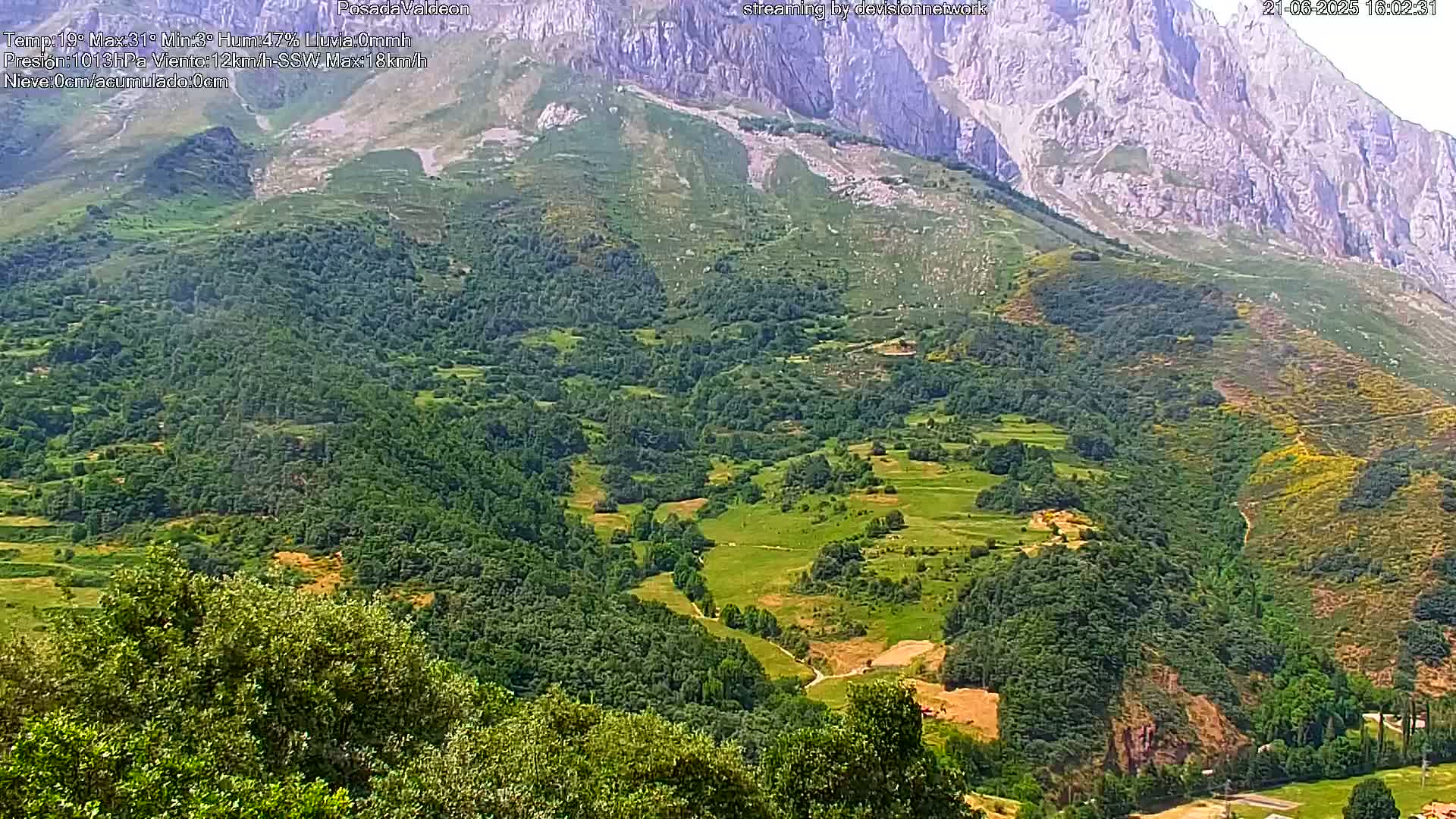 A lush green valley nestled beneath a towering, rocky mountain range on a sunny day.