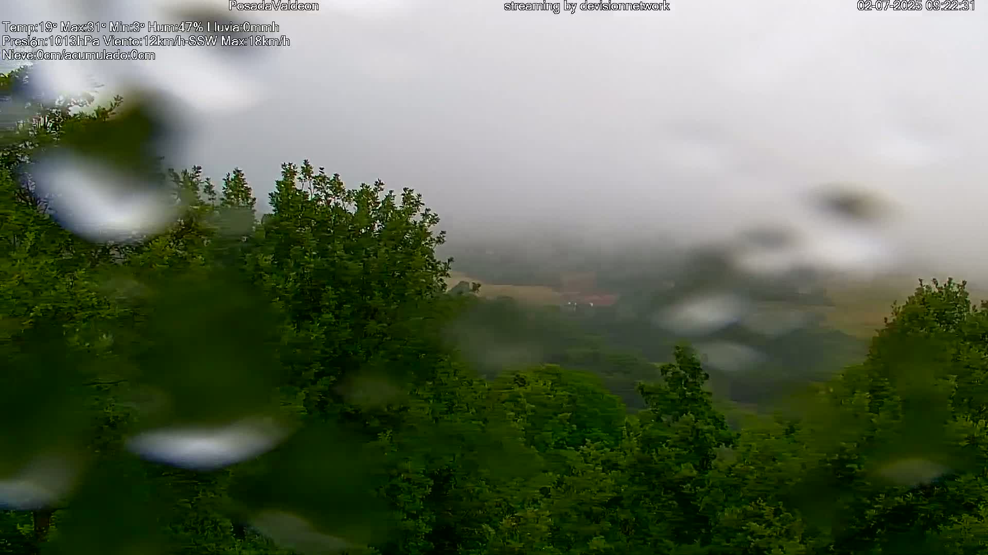 Rain falls on a view of a misty, green valley obscured by a rain-covered lens.