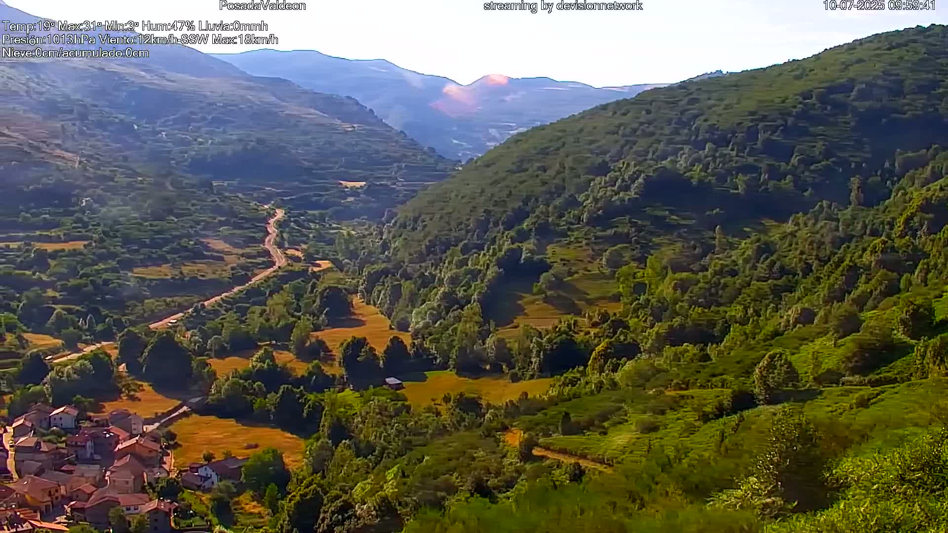 A sunny valley with a village nestled at its base, a winding road leading upward through green hills and mountains in the background.