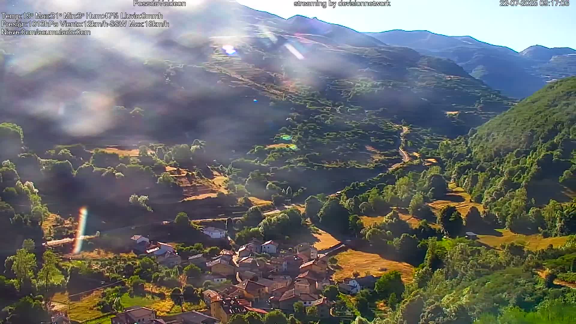 A sunlit village nestled in a verdant valley between mountains, with hazy atmospheric conditions.
