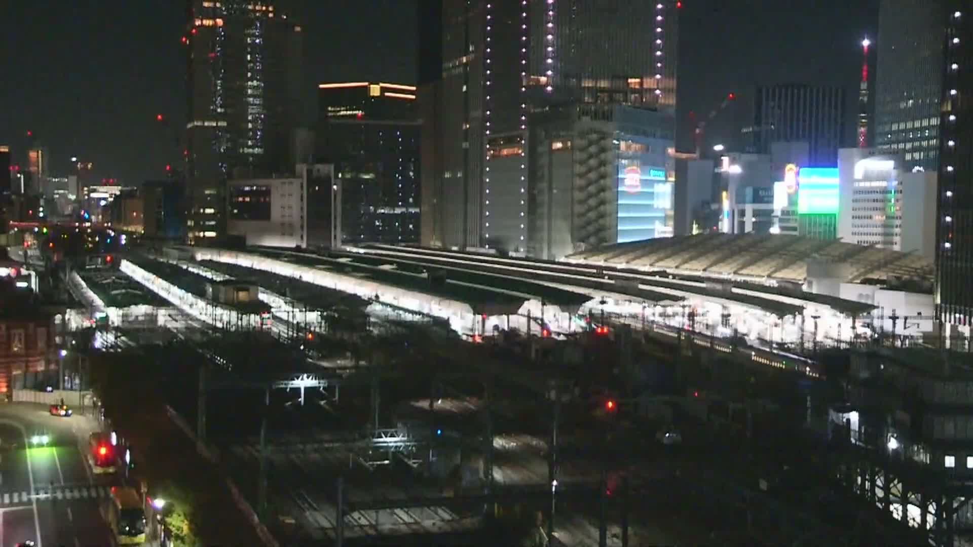 A nighttime aerial view shows a sprawling, brightly lit train station with multiple platforms and tracks, flanked by numerous illuminated skyscrapers and a busy street, all under clear skies.