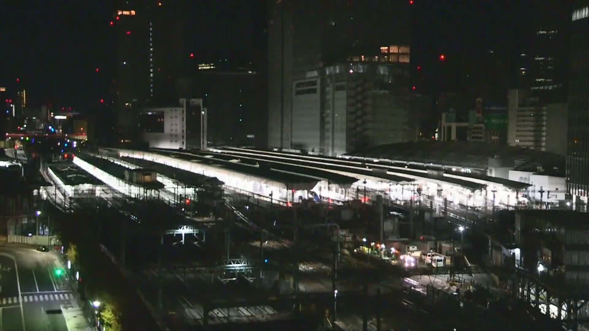 A nighttime aerial view shows a sprawling, brightly lit train station with multiple platforms and tracks, flanked by numerous illuminated skyscrapers and a busy street, all under clear skies.