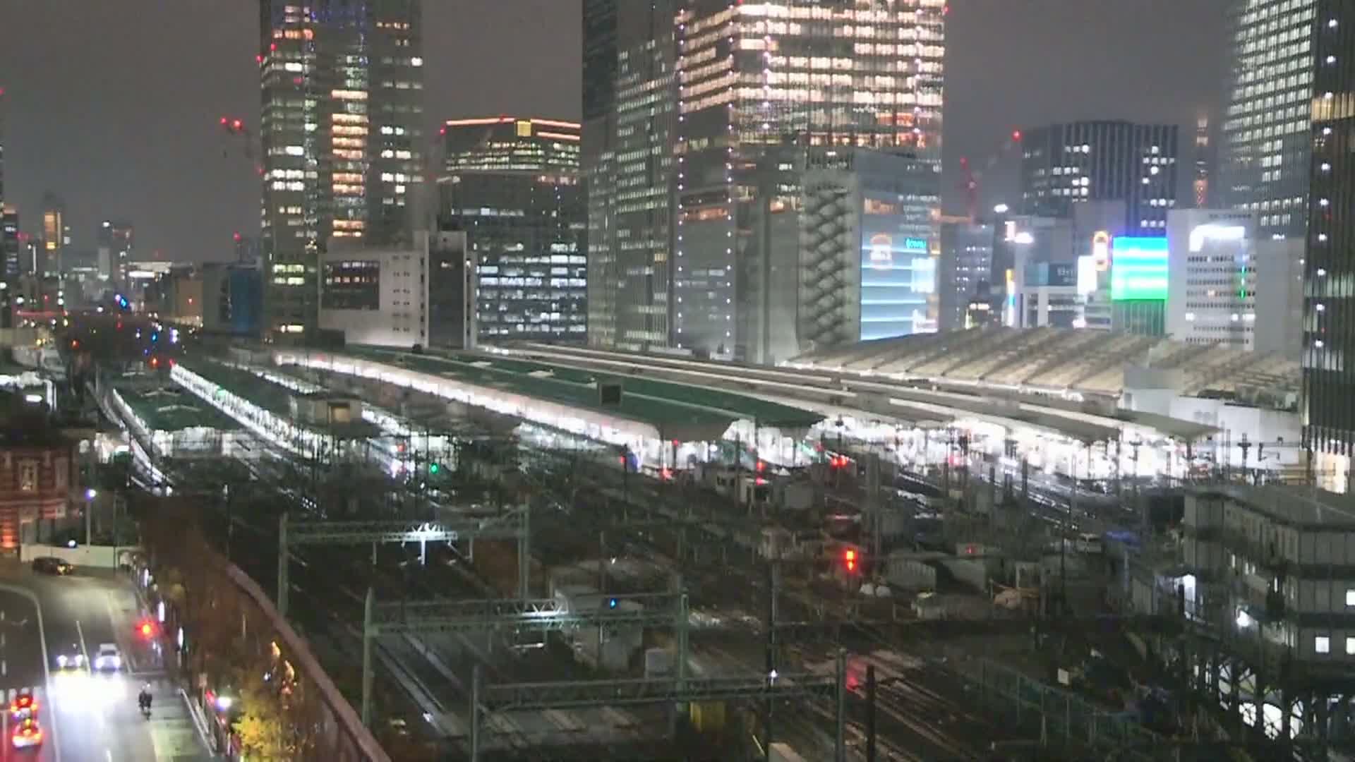 A nighttime aerial view shows a sprawling, brightly lit train station with multiple platforms and tracks, flanked by numerous illuminated skyscrapers and a busy street, all under clear skies.