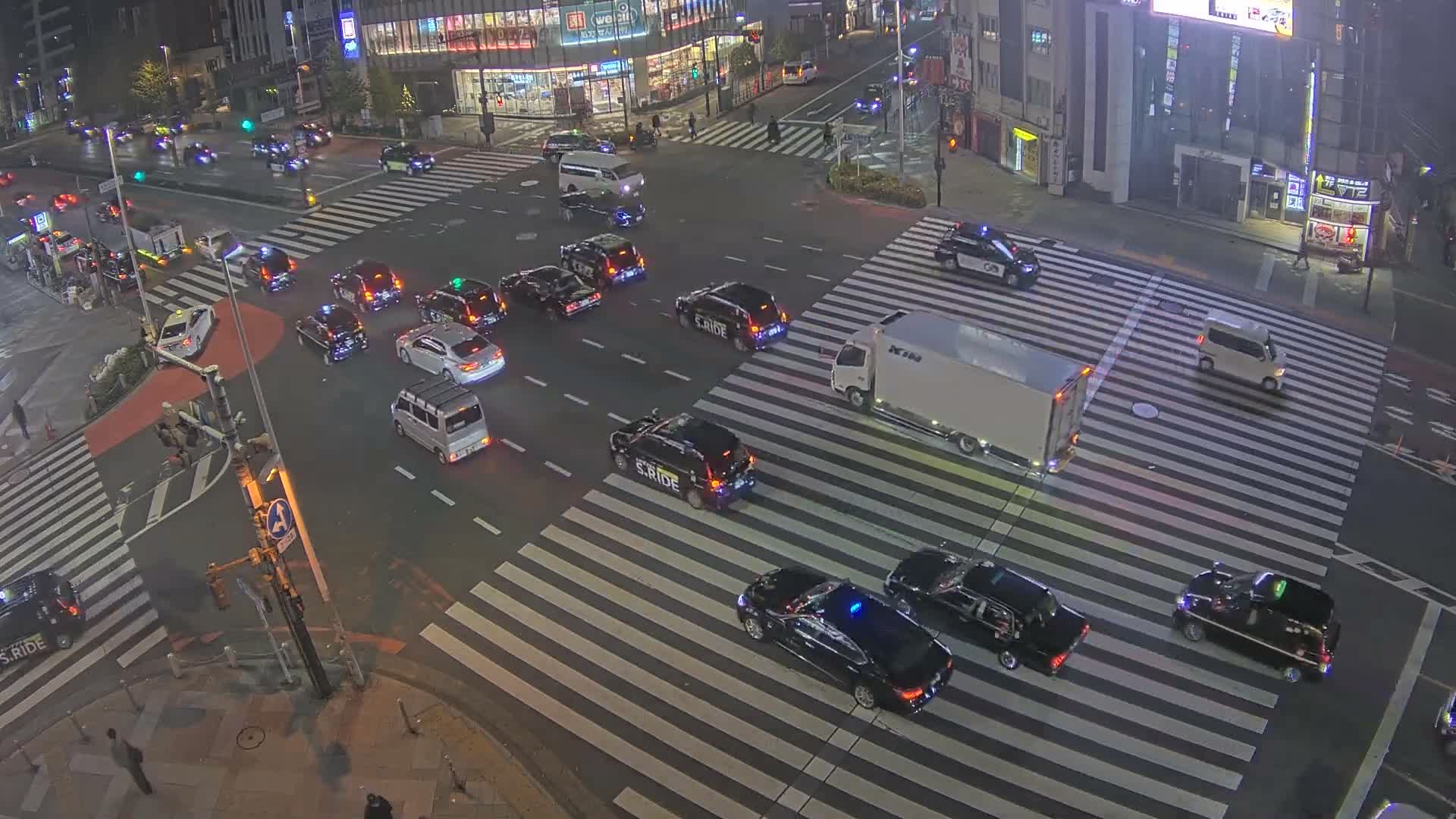 The image captures an overhead view of a bustling city intersection at night under clear weather conditions, filled with numerous vehicles, pedestrians, and illuminated buildings.