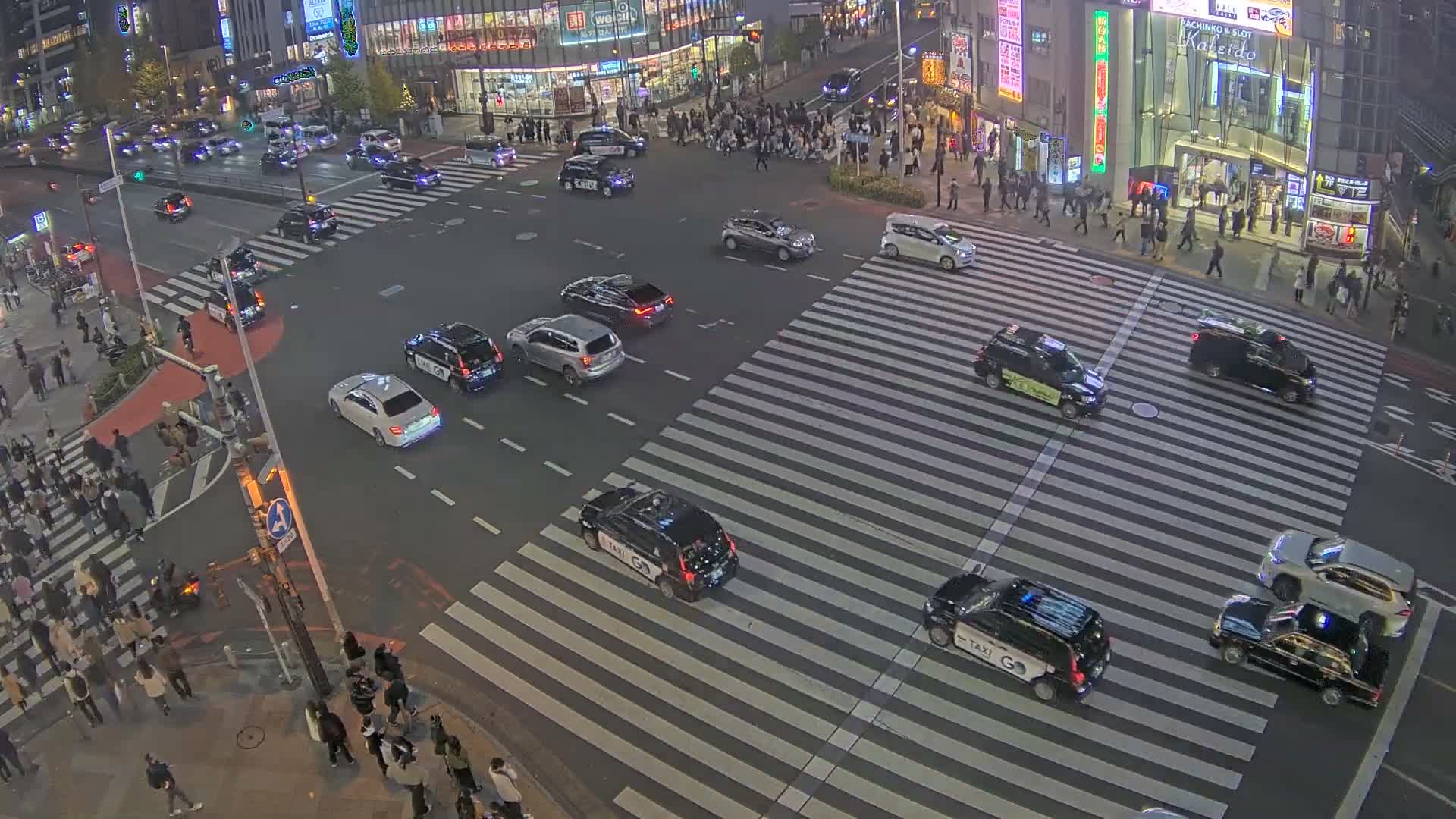 An elevated view shows a bustling city intersection at night, with a dense flow of vehicles including many taxis, and numerous pedestrians crossing well-marked crosswalks between brightly lit buildings under clear, dry weather conditions.